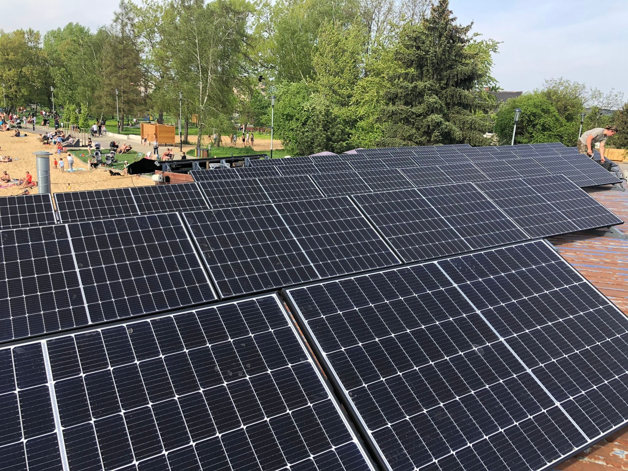 Montaż paneli fotowoltaicznych na dachu z widokiem na park miejski, gdzie widać osoby odpoczywające na plaży. Instalator w trakcie pracy. Ujęcie z góry.