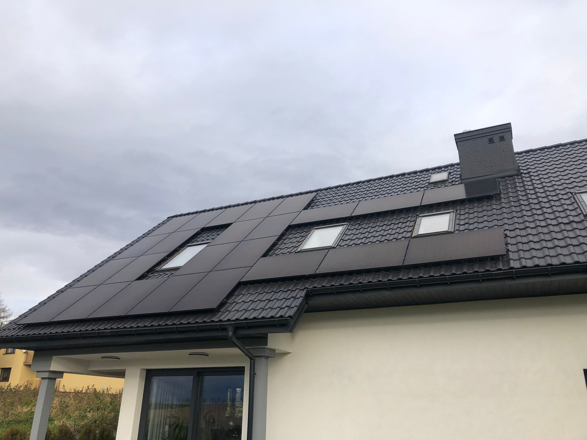 Czarny dach z panelami fotowoltaicznymi i oknami dachowymi w nowoczesnym domu jednorodzinnym, widoczne rynny i komin, pochmurne niebo w tle.