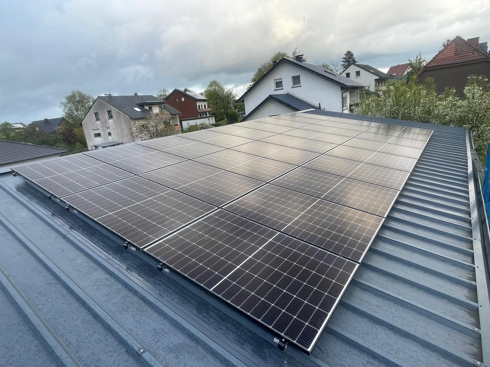 Panel słoneczny na szarym, blaszanym dachu domu jednorodzinnego w pochmurny dzień. Widok z góry, panele ułożone równo, w tle zabudowa mieszkalna.