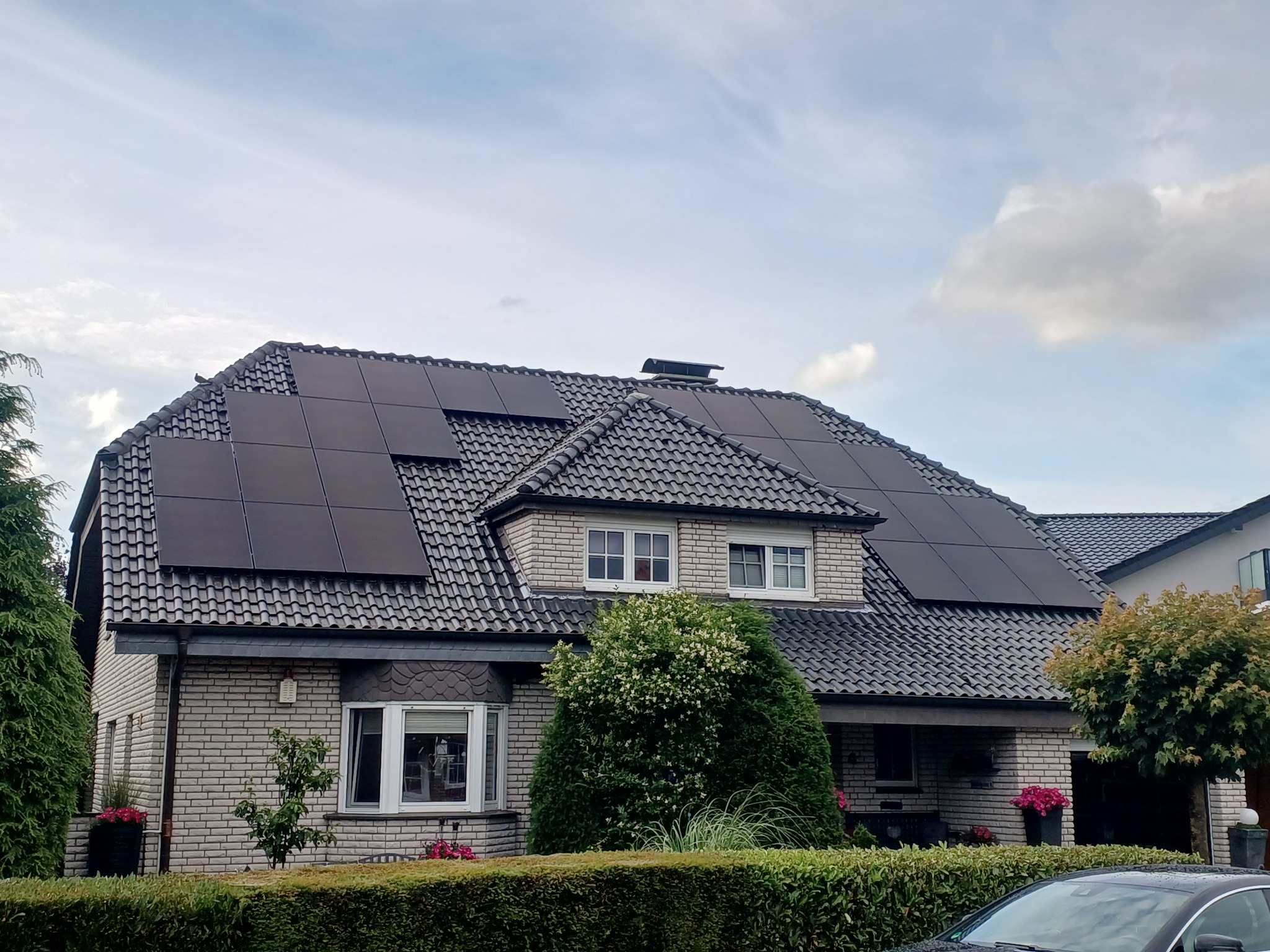 Dom z szarej cegły z panelami fotowoltaicznymi na dachu krytym szarą dachówką. Widoczne okna i zadbany ogród z krzewami.