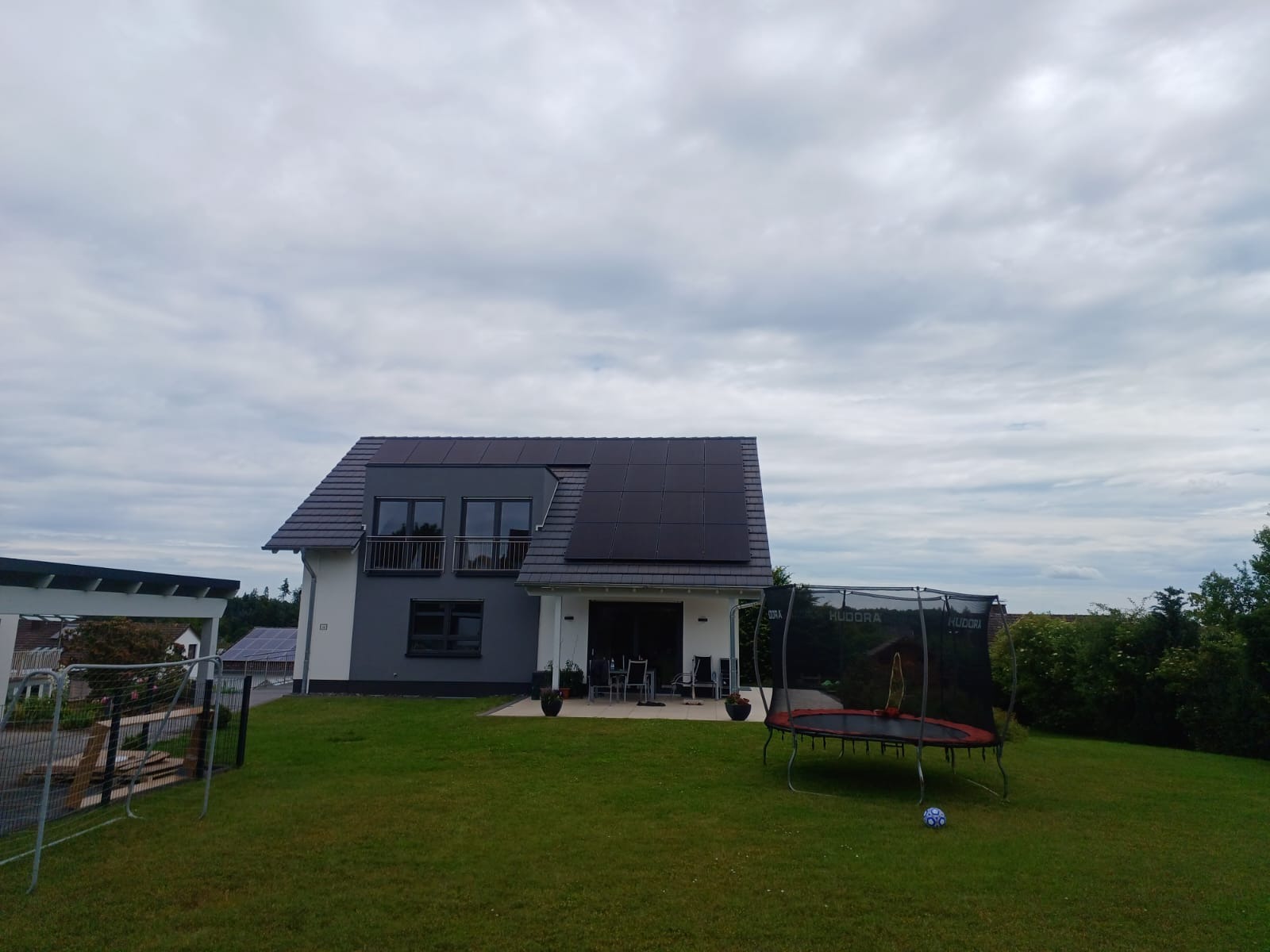 Dom jednorodzinny z panelami fotowoltaicznymi na dachu, widok z ogrodu z trampoliną i bramką piłkarską. Szaro-biała elewacja, balkon z metalową balustradą.