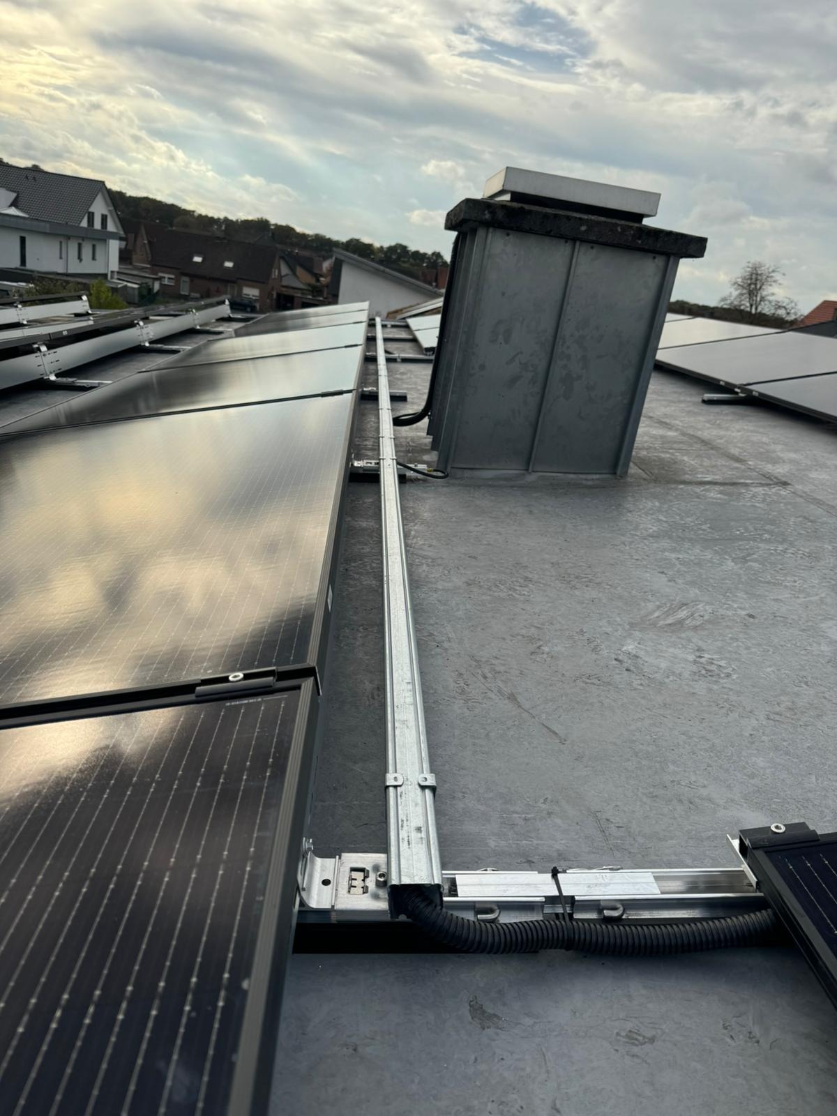 Montaż paneli fotowoltaicznych na płaskim dachu z szarym pokryciem, widoczne metalowe szyny montażowe i rura kablowa. W tle zabudowa mieszkalna i pochmurne niebo.