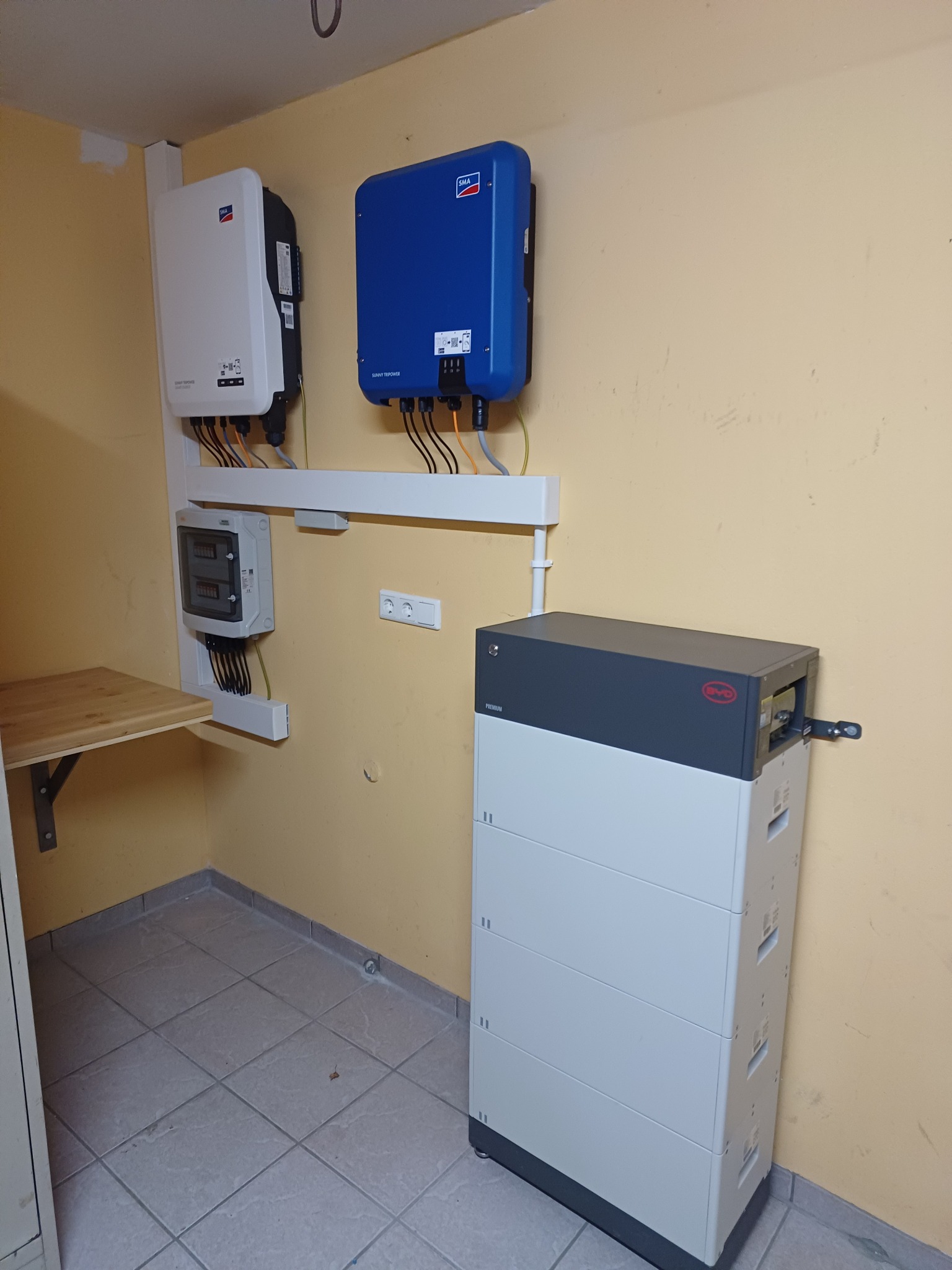 Kompletny system magazynowania energii: biały akumulator BYD, inwertery SMA (biały i niebieski) i rozdzielnia elektryczna w pomieszczeniu z żółtymi ścianami i płytkami.