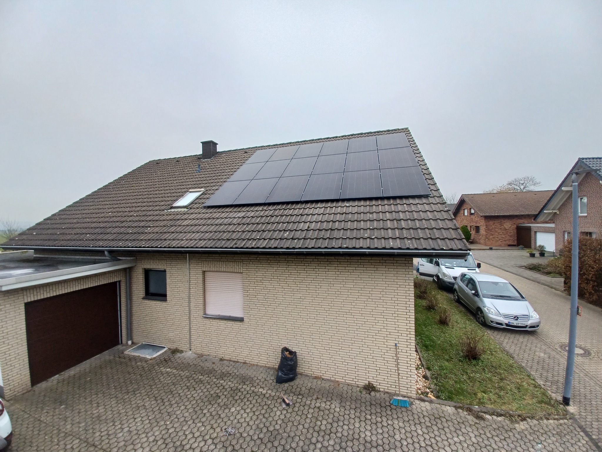 Dom z panelami słonecznymi na brązowym dachu, widok z góry. Na podjeździe zaparkowane dwa samochody i widać czarny worek na śmieci.