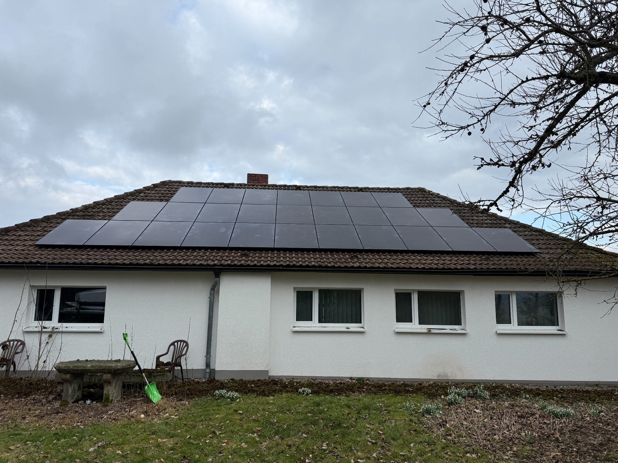 Dom z panelami fotowoltaicznymi na dachu z brązowej dachówki, widoczne okna i elementy ogrodu. Pochmurne niebo w tle.