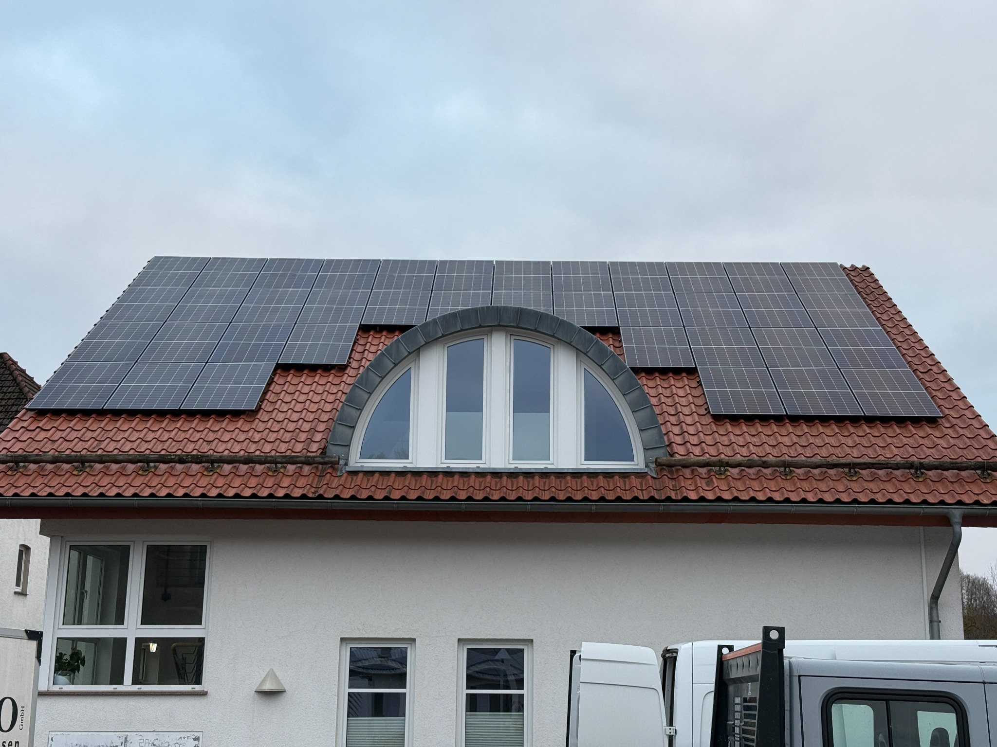 Panele fotowoltaiczne na dachu domu z czerwoną dachówką, widoczne okna dachowe i fragmenty zaparkowanych pojazdów. Pochmurne niebo w tle.