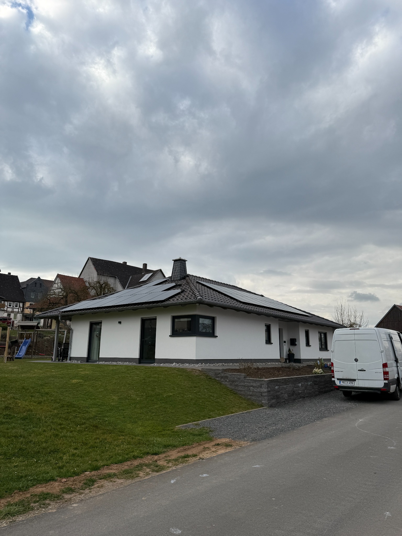 Biały dom jednorodzinny z panelami słonecznymi na dachu, na tle pochmurnego nieba. Z prawej strony zaparkowany biały bus. Widok z poziomu ulicy.