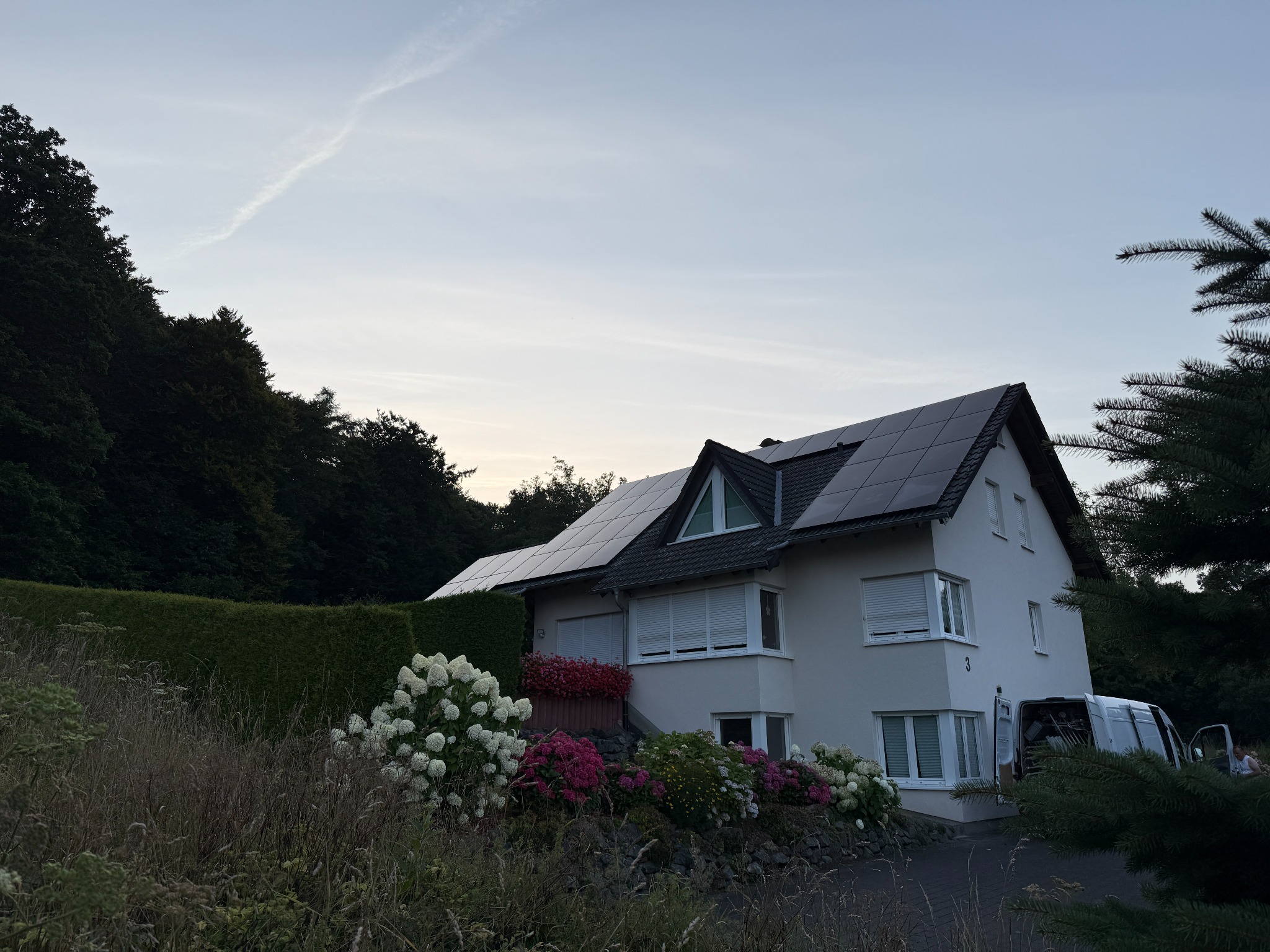 Biały dom z panelami słonecznymi na dachu, otoczony zielenią i kwiatami. Widoczny van z otwartymi drzwiami. Zachmurzone niebo.