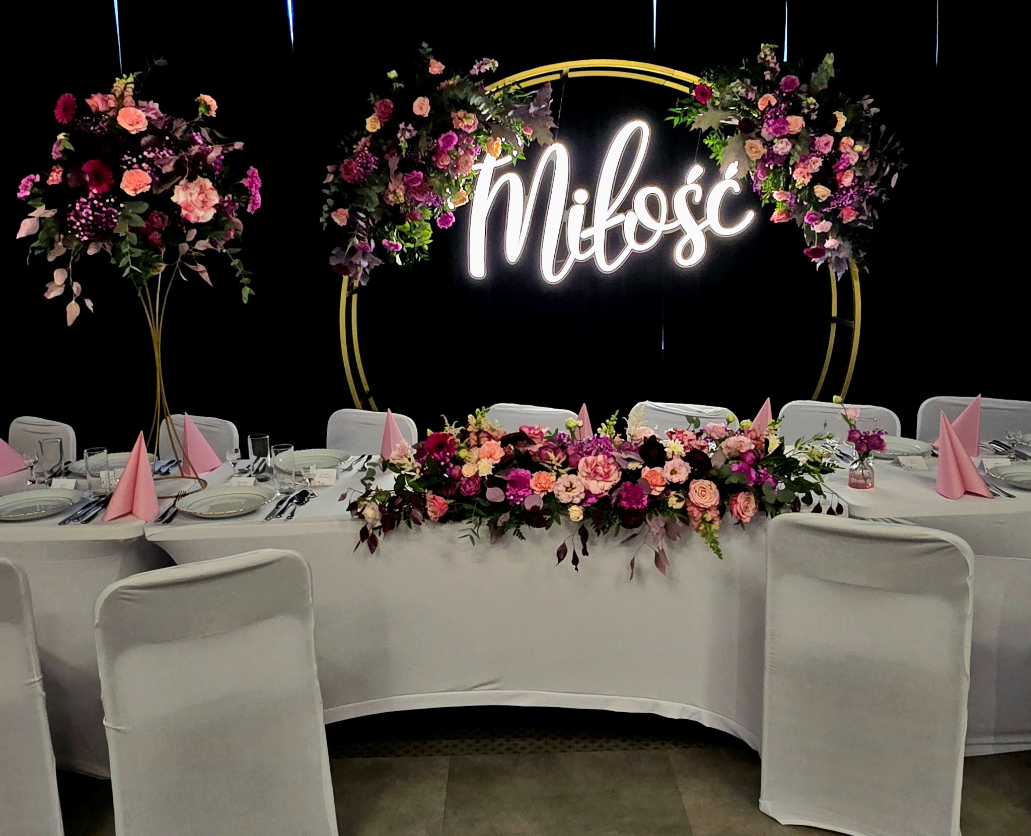 Uroczysty stół weselny z dekoracjami kwiatowymi w odcieniach różu i fioletu, podświetlonym napisem 'Miłość' i elegancką zastawą.