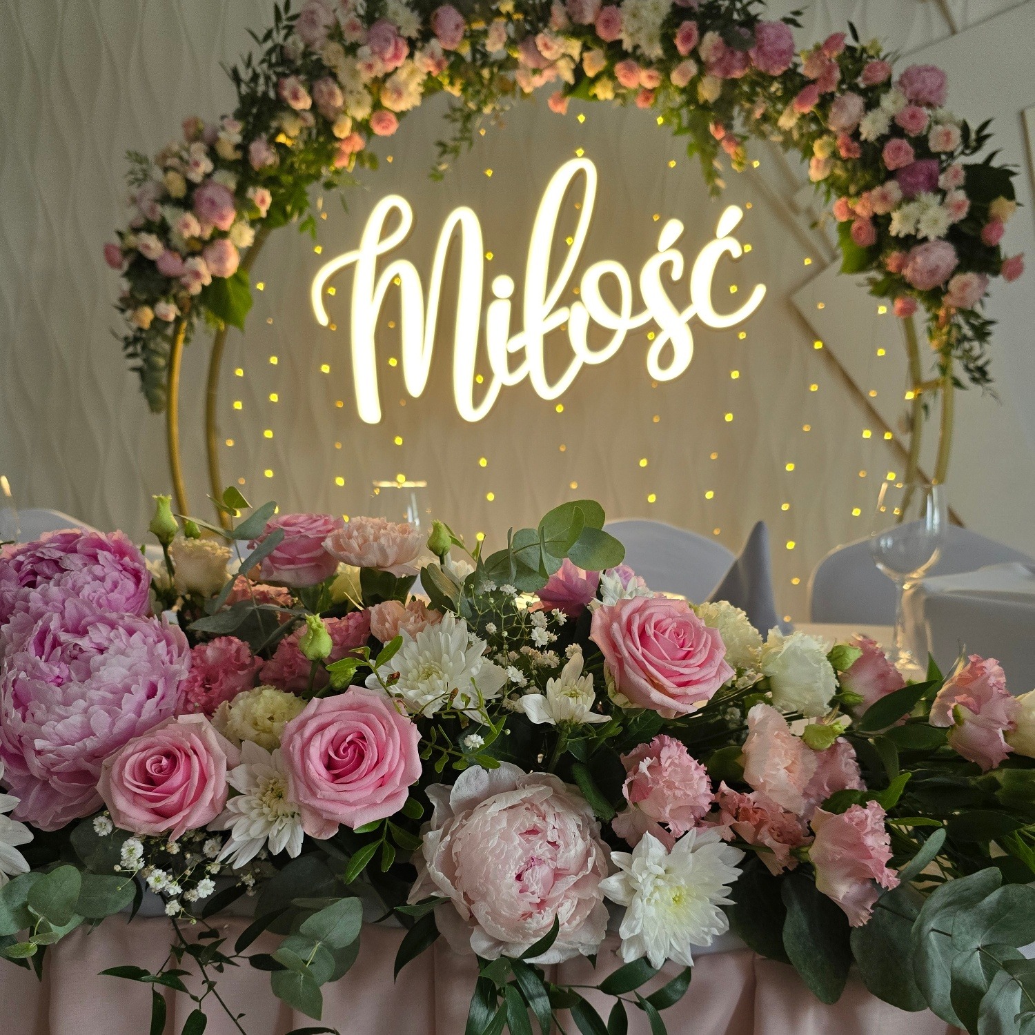 Dekoracja stołu weselnego w Łodzi: kwiaty różowe i białe, neon 'Miłość' na złotym stelażu, subtelne oświetlenie. Romantyczny wystrój sali.