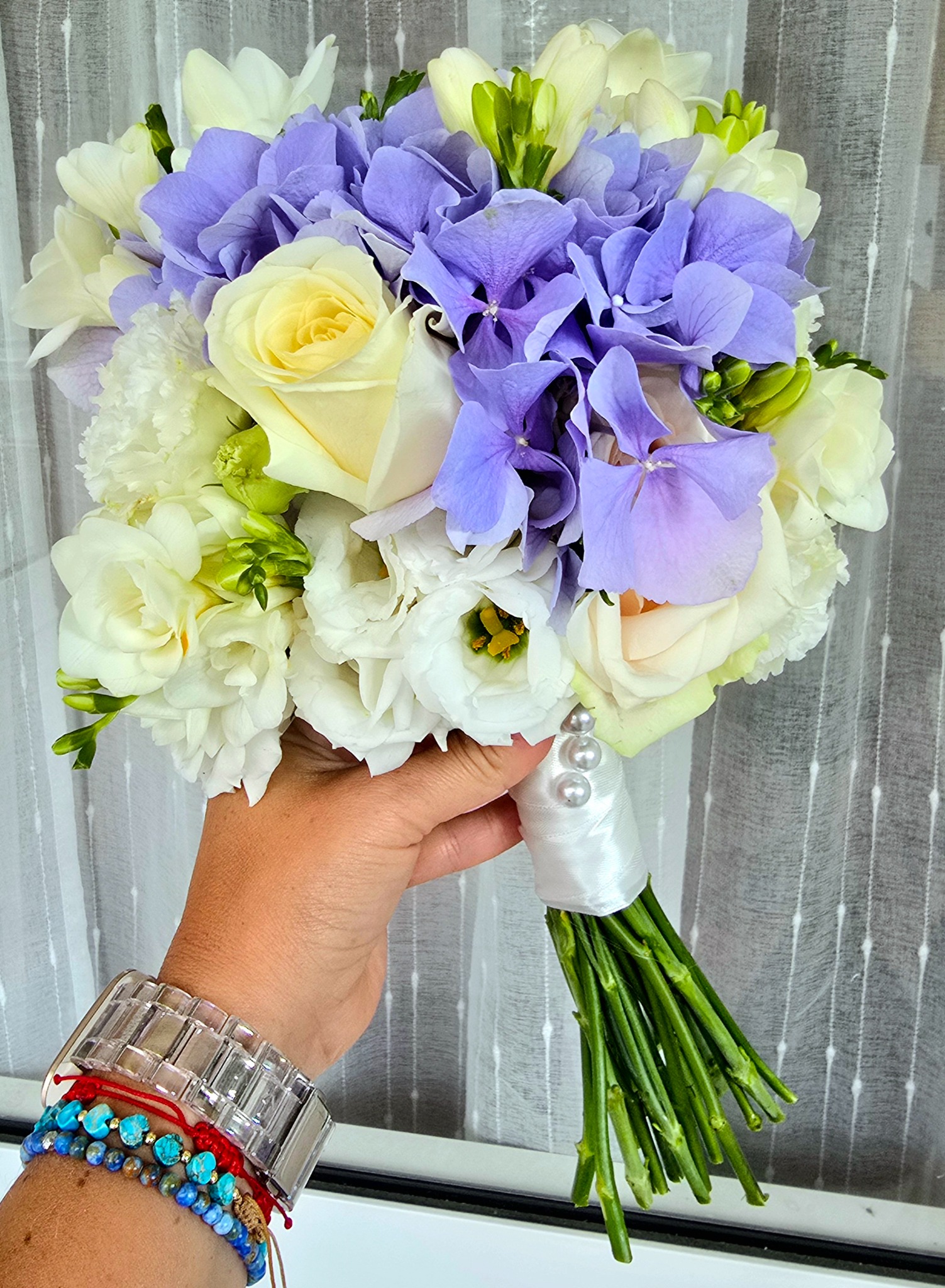 Biało-fioletowy bukiet ślubny z róż i hortensji, trzymany w dłoni z bransoletkami na tle białej, wzorzystej firanki. Elegancki i świeży.