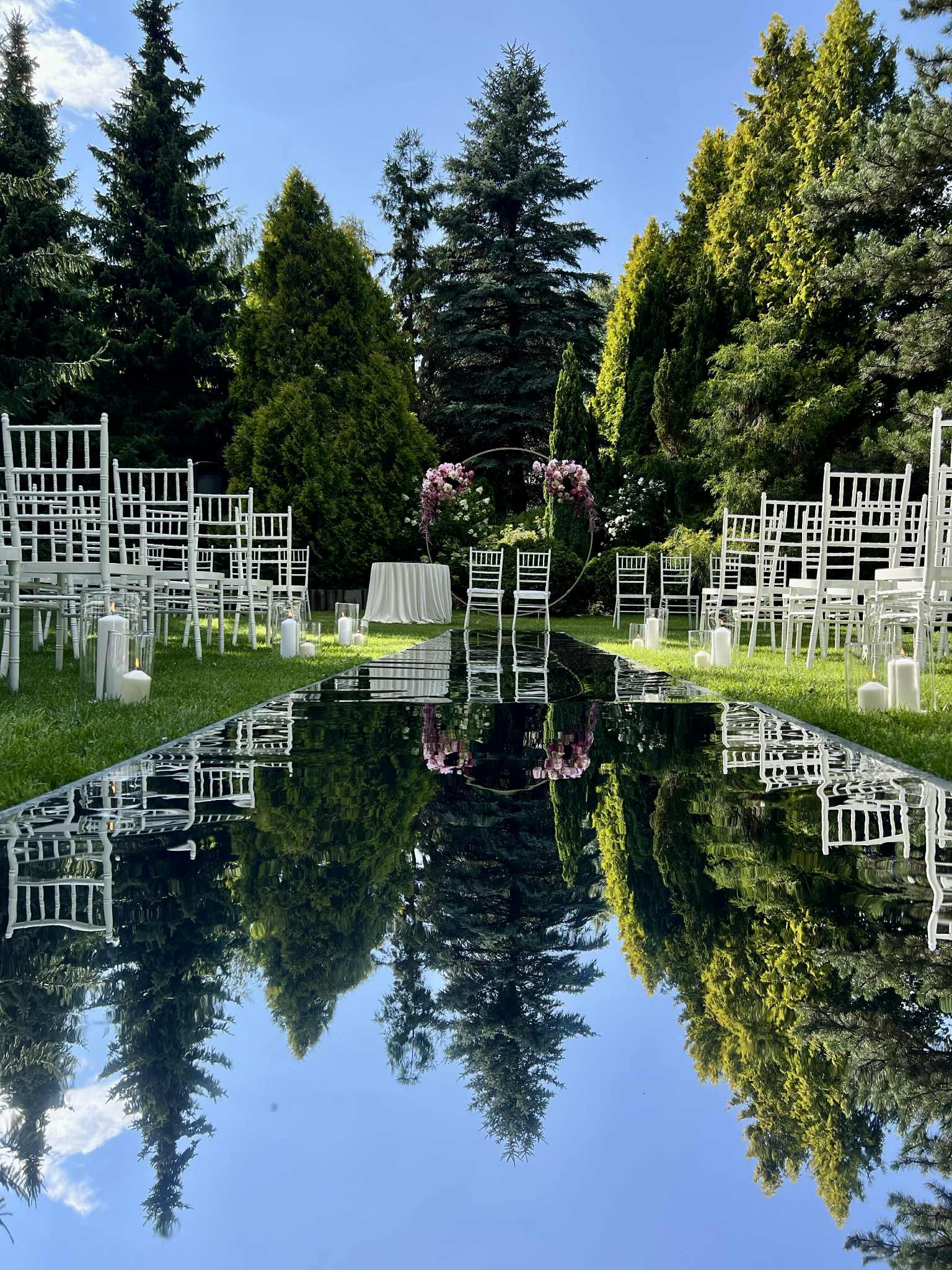 Uroczysta ścieżka z lustrzaną podłogą, otoczona białymi krzesłami i świecami, prowadząca do kwiatowej bramy na tle zieleni, gotowa na ceremonię ślubną.
