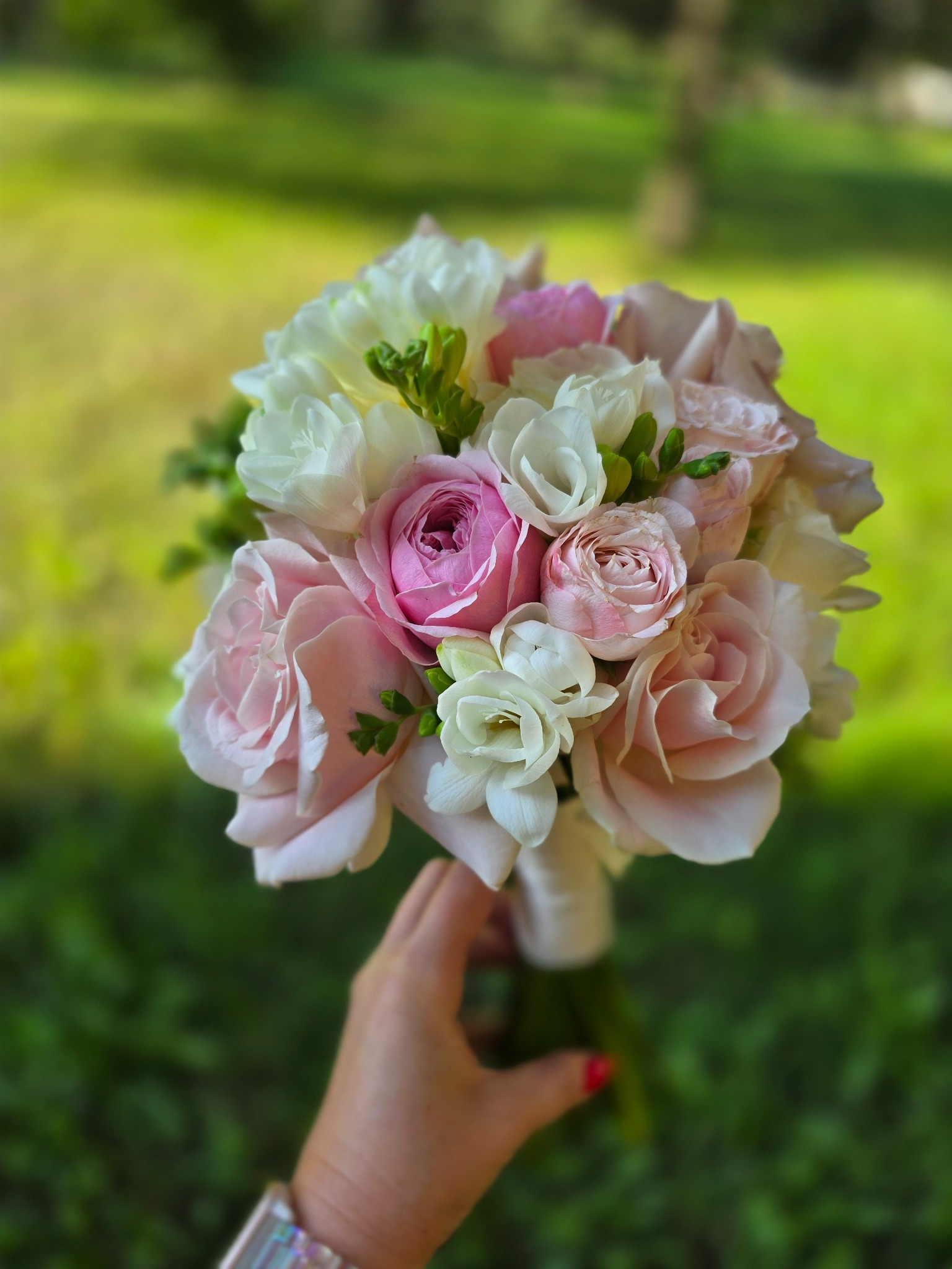 Delikatny bukiet ślubny z różowych i białych róż trzymany w dłoni na tle rozmytej zieleni. Widoczny fragment zegarka na nadgarstku.