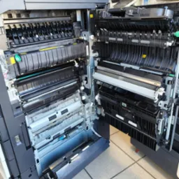 Otwarta kserokopiarka A3 ukazująca wewnętrzne mechanizmy, rolki i podajniki papieru; widoczne elementy konstrukcyjne i ostrzeżenia na obudowie.