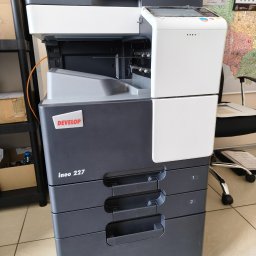 R-TECH ŁUKASZ ROSOWSKI - Kserokopiarka Develop ineo 227 w biurze, z trzema szufladami na papier i panelem sterowania. Widoczny kalendarz na ścianie i krzesło biurowe w tle.