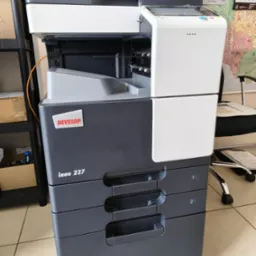 Kserokopiarka Develop ineo 227 w biurze, z trzema szufladami na papier i panelem sterowania. Widoczny kalendarz na ścianie i krzesło biurowe w tle.