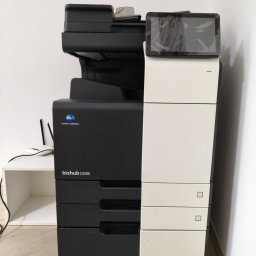 R-TECH ŁUKASZ ROSOWSKI - Stojąca kserokopiarka Konica Minolta bizhub C250i, widok z przodu, obok biały regał z routerem. Ekran dotykowy kserokopiarki lekko zabrudzony.