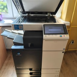 R-TECH ŁUKASZ ROSOWSKI - Kserokopiarka Konica Minolta bizhub 364e z otwartą klapą skanera, widoczny ekran dotykowy z menu logowania. Urządzenie stoi na drewnianej podłodze przy żółtej ścianie.