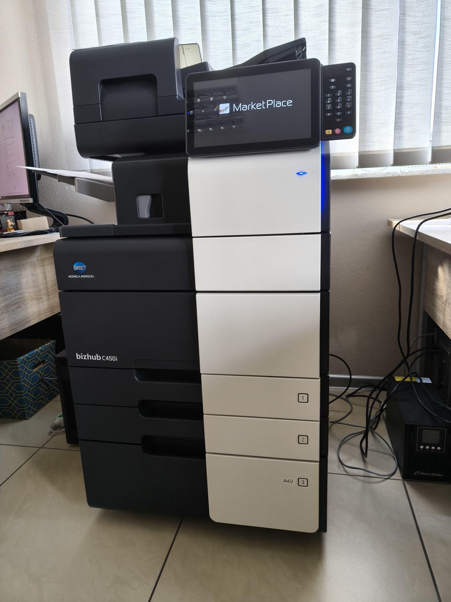 Wielofunkcyjna kserokopiarka Konica Minolta bizhub C450i z panelem dotykowym Market Place, widoczna w biurze, zasilacz awaryjny PowerWalker obok.