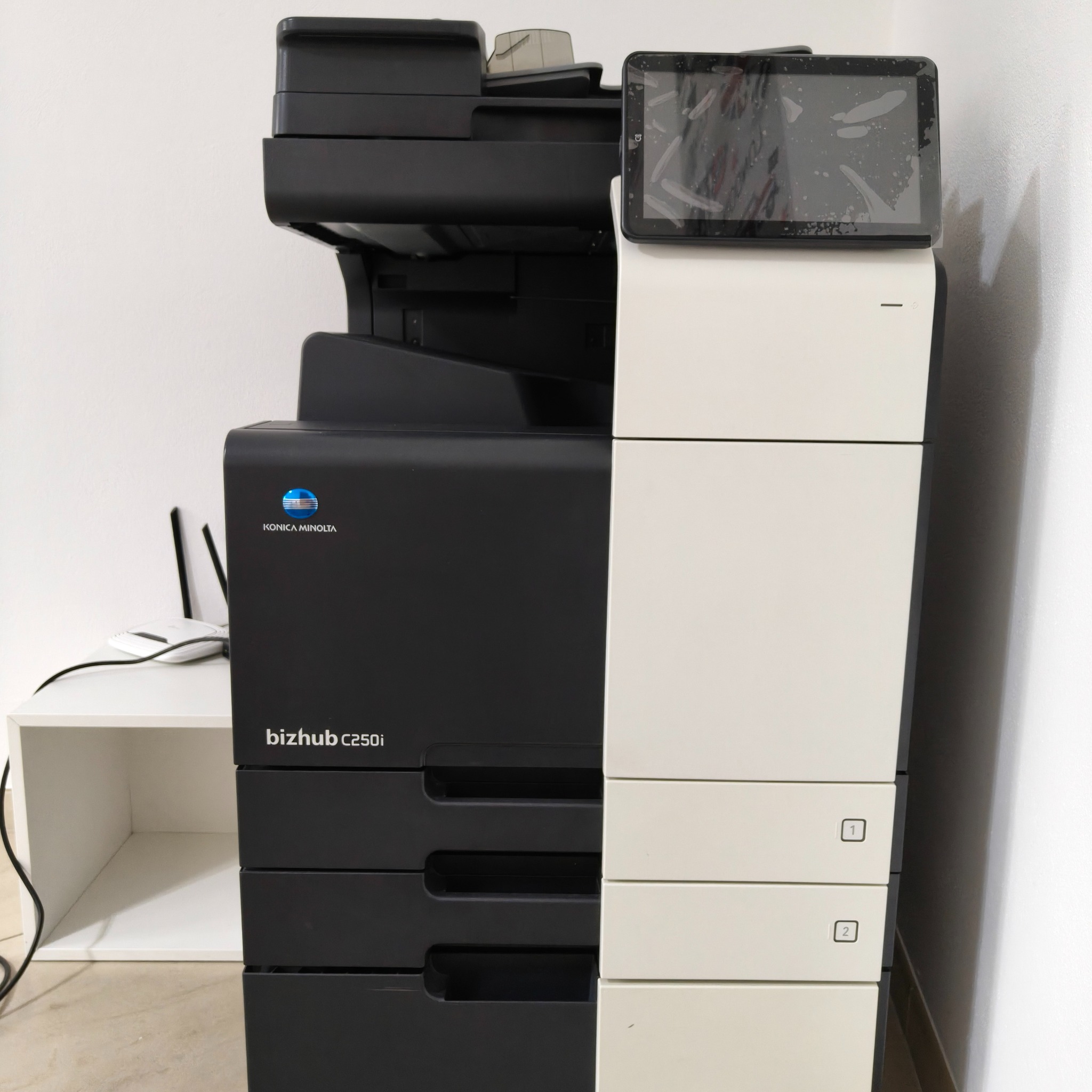 Stojąca kserokopiarka Konica Minolta bizhub C250i, widok z przodu, obok biały regał z routerem. Ekran dotykowy kserokopiarki lekko zabrudzony.