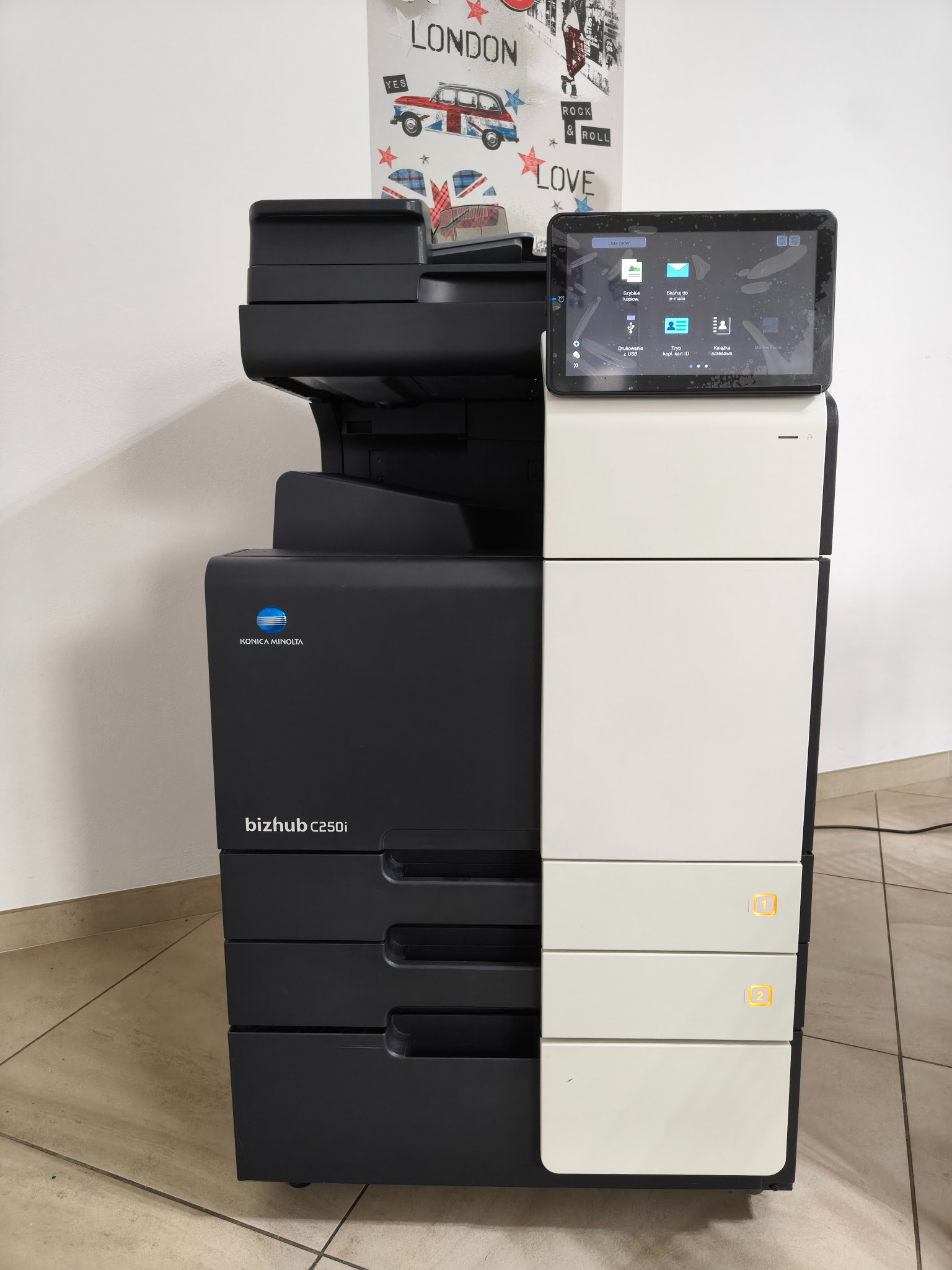 Czarno-biała kserokopiarka Konica Minolta bizhub C250i z dotykowym ekranem, na tle ściany z plakatem o tematyce londyńskiej, widok z przodu, lekko z dołu.