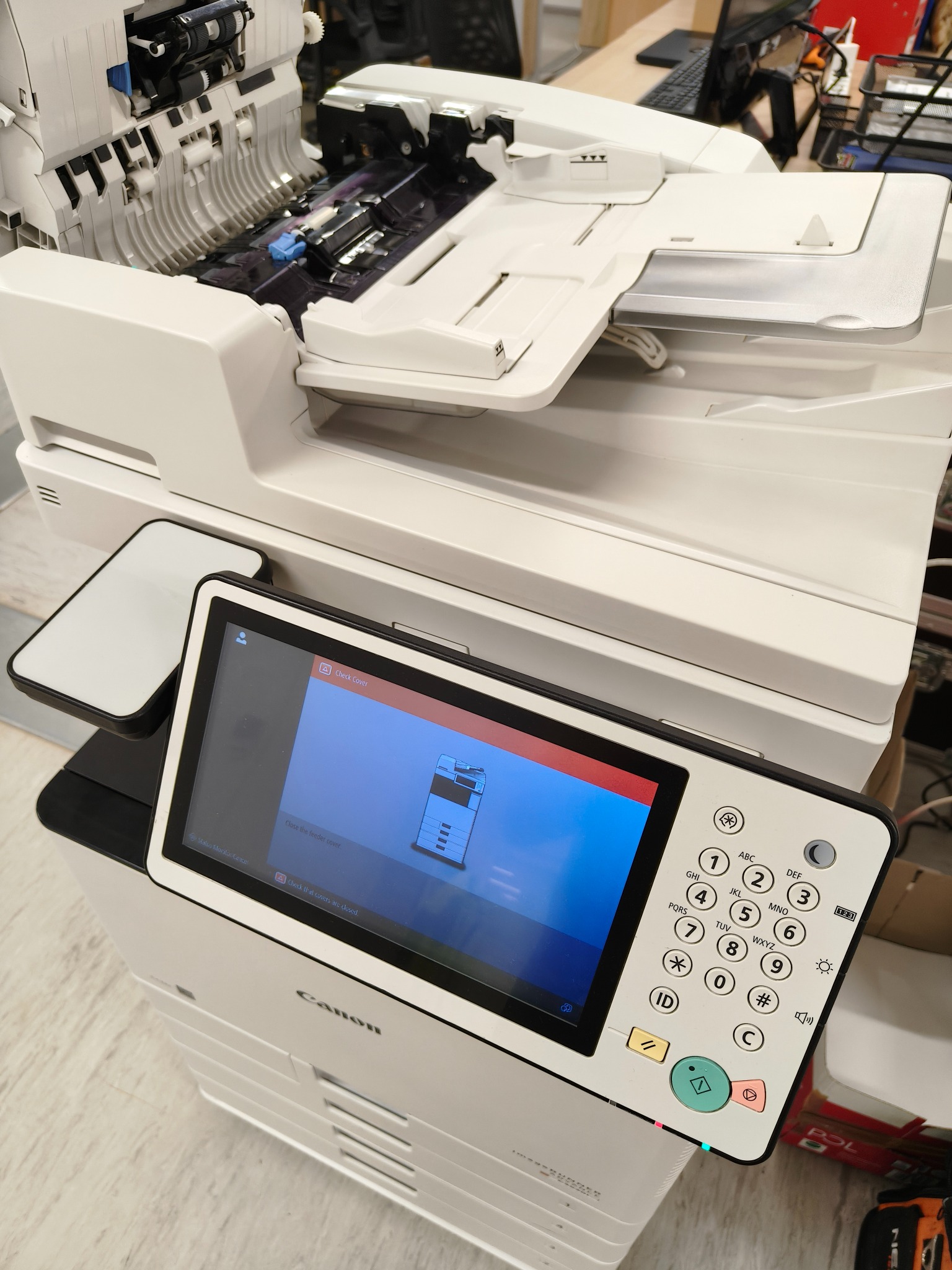 Biała kserokopiarka Canon w trakcie naprawy, z otwartą obudową i komunikatem o błędzie na ekranie dotykowym. Widoczna klawiatura urządzenia i elementy wewnętrzne.