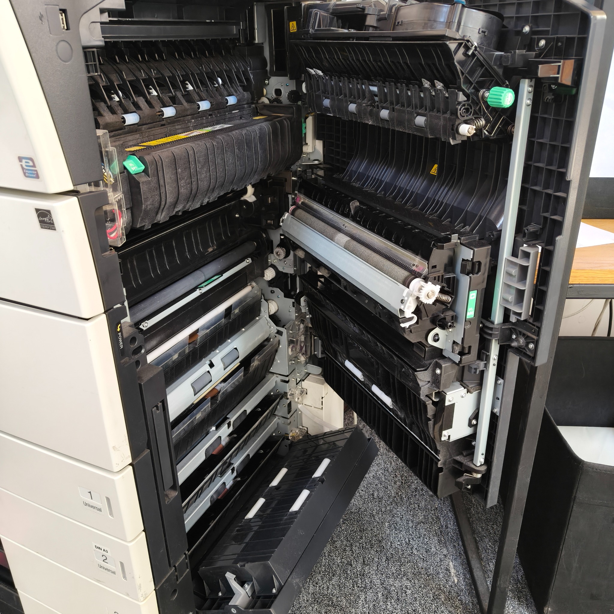 Otwarta kserokopiarka wielkoformatowa w trakcie serwisu, widoczne wewnętrzne mechanizmy i podajniki papieru. Detal urządzenia biurowego podczas naprawy.