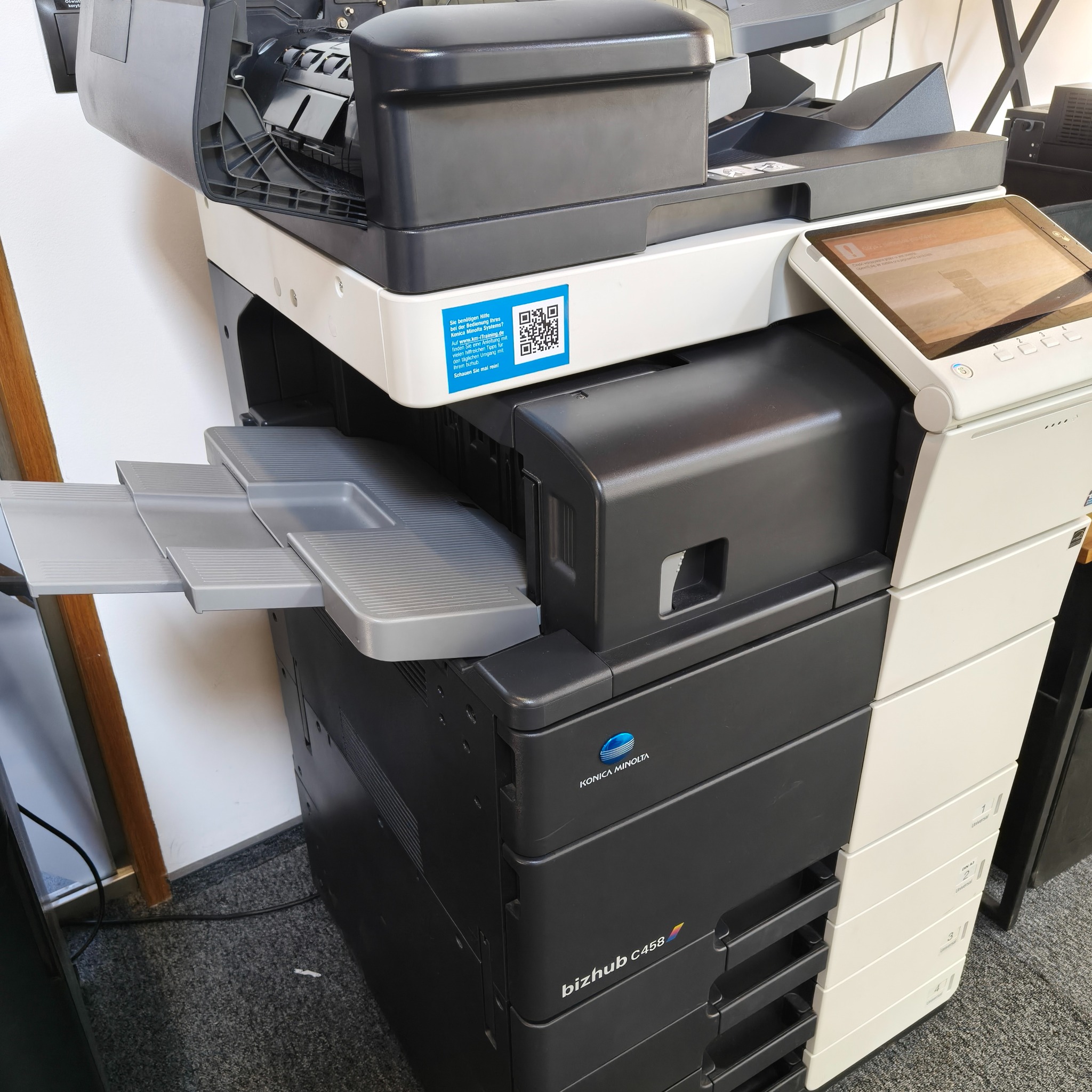 Wielofunkcyjna kserokopiarka Konica Minolta bizhub C458 z wysuniętą szufladą na papier i panelem dotykowym, widoczna naklejka z kodem QR. Urządzenie stoi na dywanie.