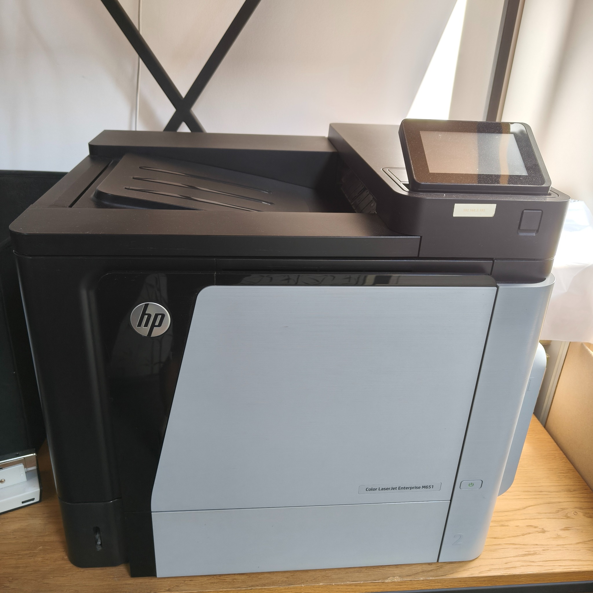 Czarno-szara kserokopiarka HP Color LaserJet Enterprise M651 z panelem dotykowym i logo HP, stojąca na drewnianej półce. Widoczny fragment otoczenia.