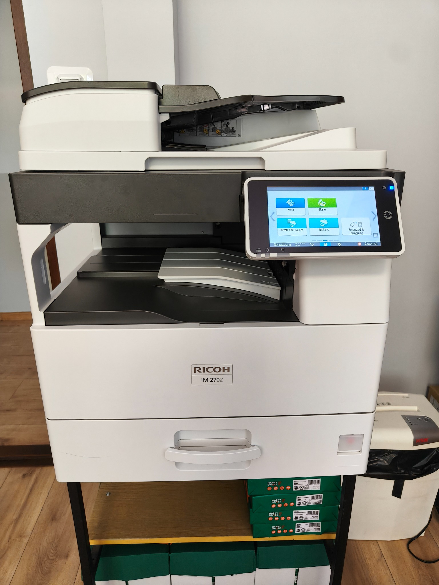 Nowoczesna, biała kserokopiarka Ricoh IM 2702 z panelem dotykowym prezentującym opcje kopiowania i skanowania, umieszczona na półce z zapasem papieru. Widoczny również niszczarka.