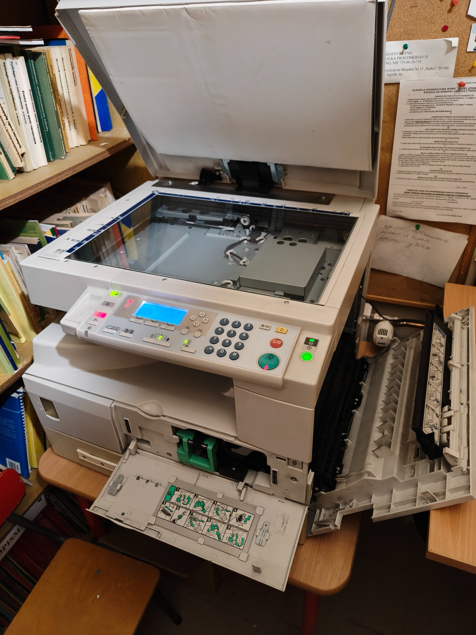 Otwarta kserokopiarka biurowa z widocznymi elementami wewnętrznymi, obok półka z książkami i dokumenty. Sprzęt prawdopodobnie w trakcie naprawy lub konserwacji.