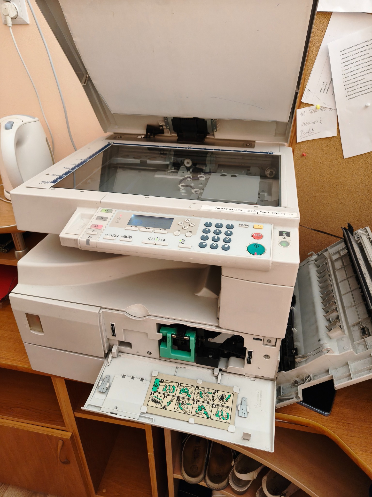 Otwarty skaner kserokopiarki z widocznym wnętrzem, obok tablica korkowa z notatkami. Widoczny panel sterowania i instrukcja obsługi w otwartej szufladzie. Sprzęt biurowy w trakcie serwisu.