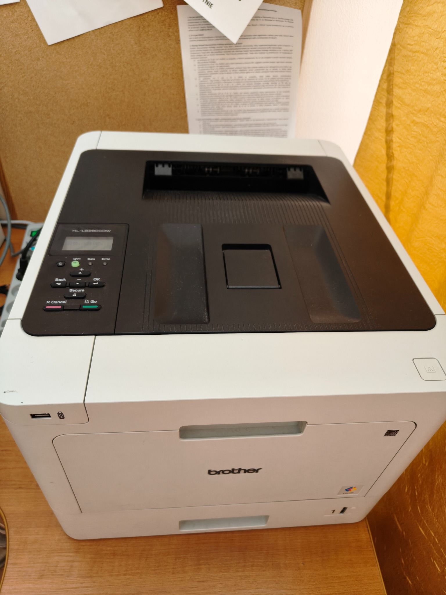 Biała drukarka Brother HL-L8260CDW na biurku, widoczny panel sterowania z przyciskami, logo Brother i wlot papieru. W tle dokumenty i żółta zasłona.