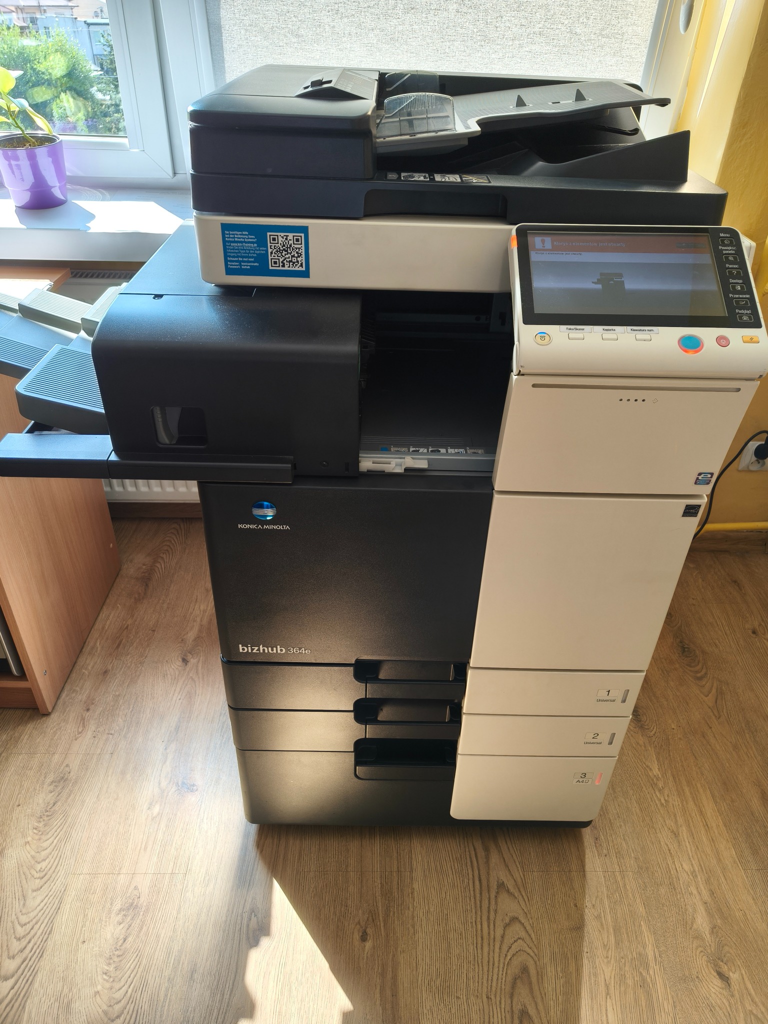 Wielofunkcyjna kserokopiarka Konica Minolta bizhub 364e, stojąca na drewnianej podłodze w pobliżu okna z rośliną. Widoczny panel sterowania z komunikatem o błędzie.