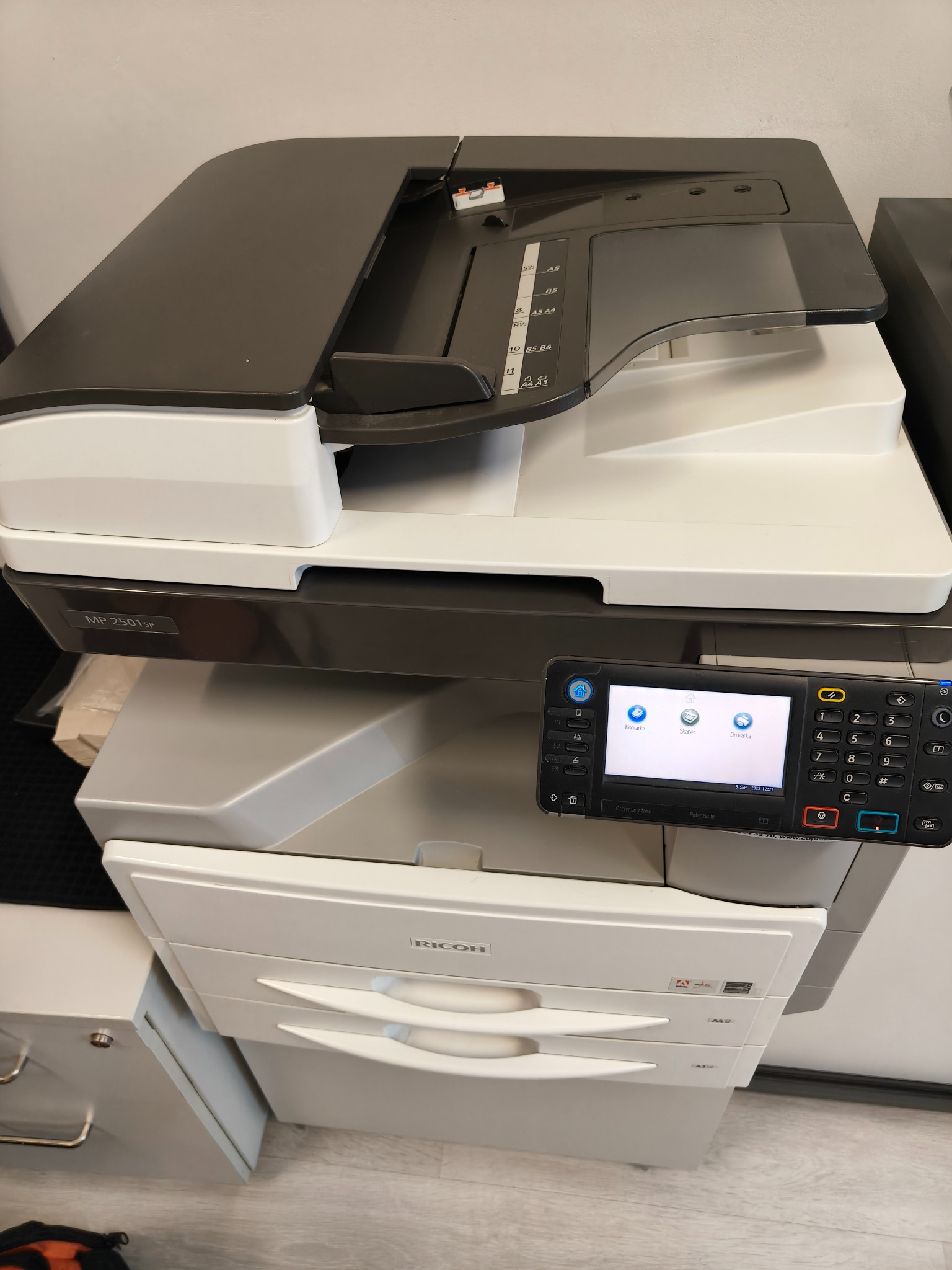 Wielofunkcyjne urządzenie Ricoh MP 2501SP z panelem dotykowym, widoczne opcje kopiowania, skanowania i drukowania. Widoczne podajniki papieru i formaty A3/A4.