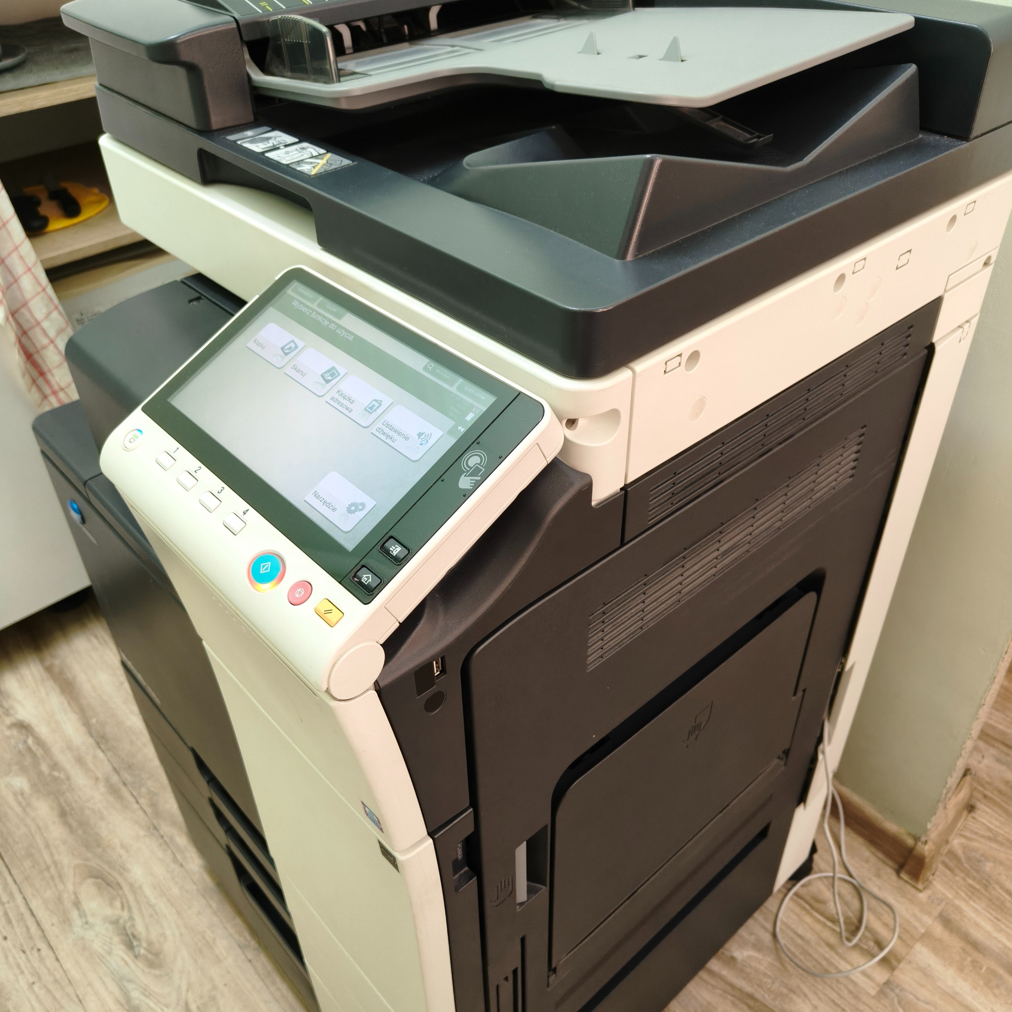 Wielofunkcyjna kserokopiarka biurowa z panelem dotykowym, widoczny interfejs użytkownika. Urządzenie w kolorze czarno-białym, ustawione na podłodze w biurze.