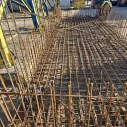 Zbrojenie fundamentu z widoczną siatką stalową i wystającymi prętami, przygotowane do zalania betonem na placu budowy. Widoczny dźwig i maszyny budowlane.