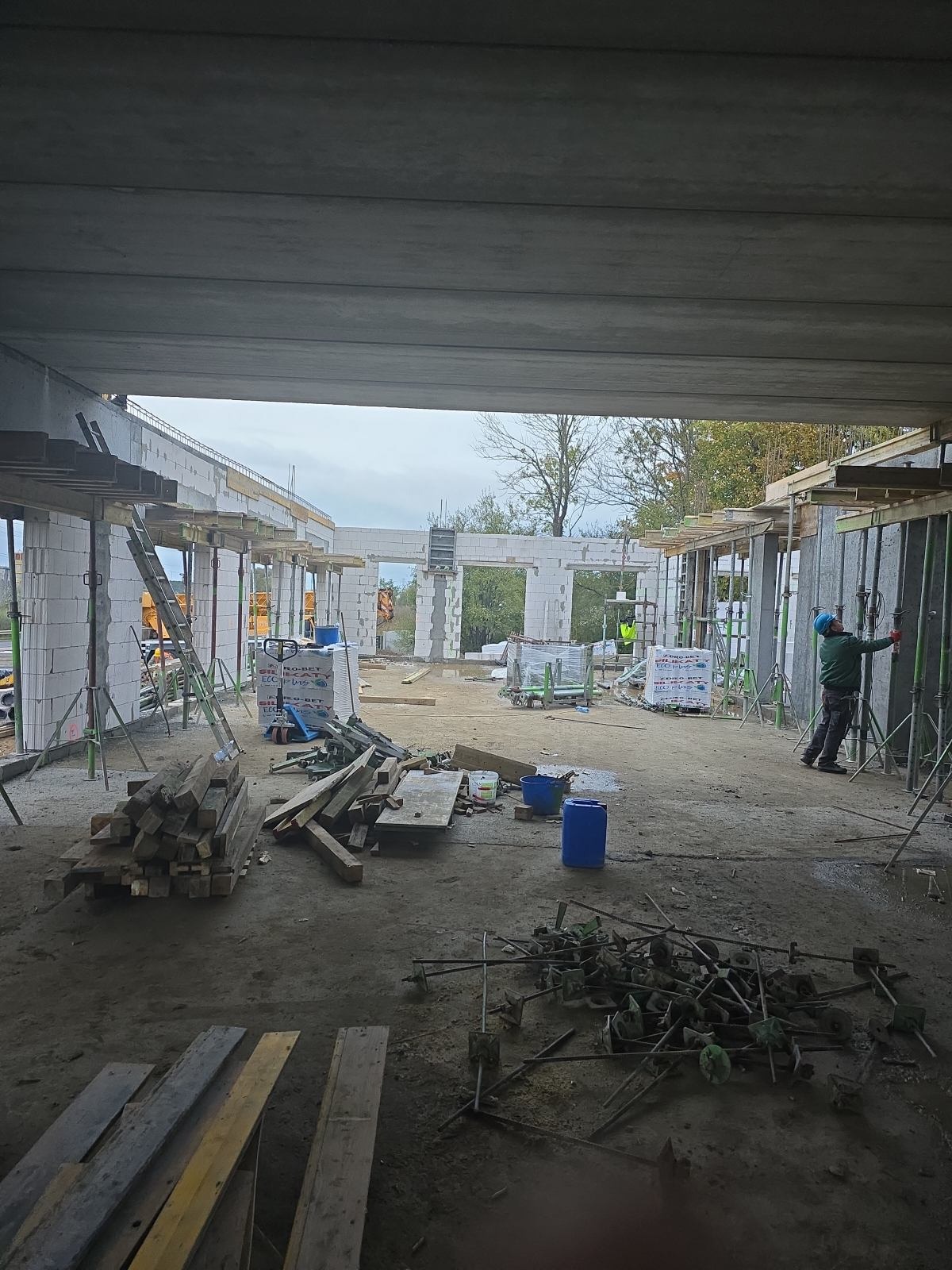 Budowa domu z betonu komórkowego: ściany wzniesione, widoczne podpory i materiały budowlane. Pracownik w kasku reguluje szalunek. W tle drzewa.