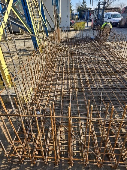 Zbrojenie fundamentu z widoczną siatką stalową i wystającymi prętami, przygotowane do zalania betonem na placu budowy. Widoczny dźwig i maszyny budowlane.