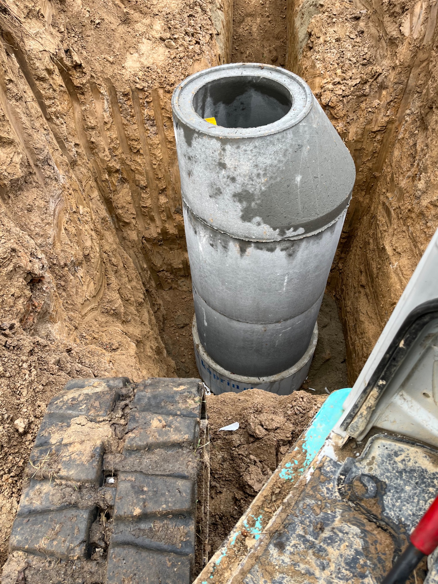 Betonowe kręgi w wykopie, widoczne ślady pracy koparki, ziemia po bokach. Elementy instalacji kanalizacyjnej w trakcie montażu.