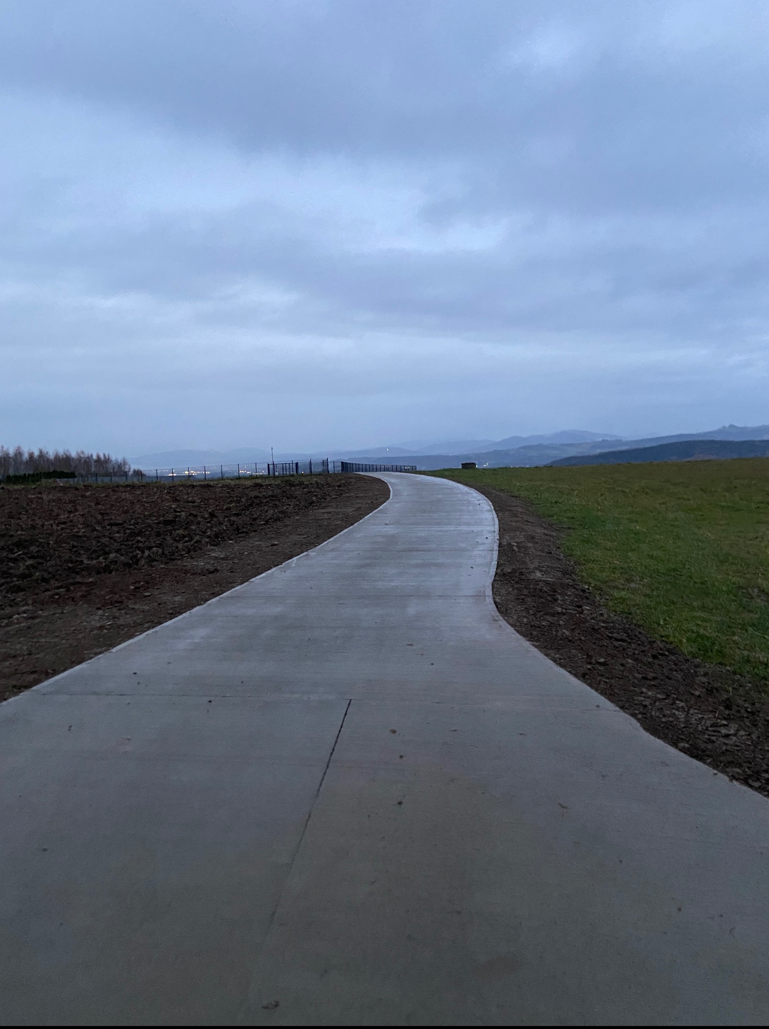 Betonowa ścieżka wijąca się przez teren z ziemią i trawą, pod pochmurnym niebem. W tle widoczne drzewa i odległe wzgórza.