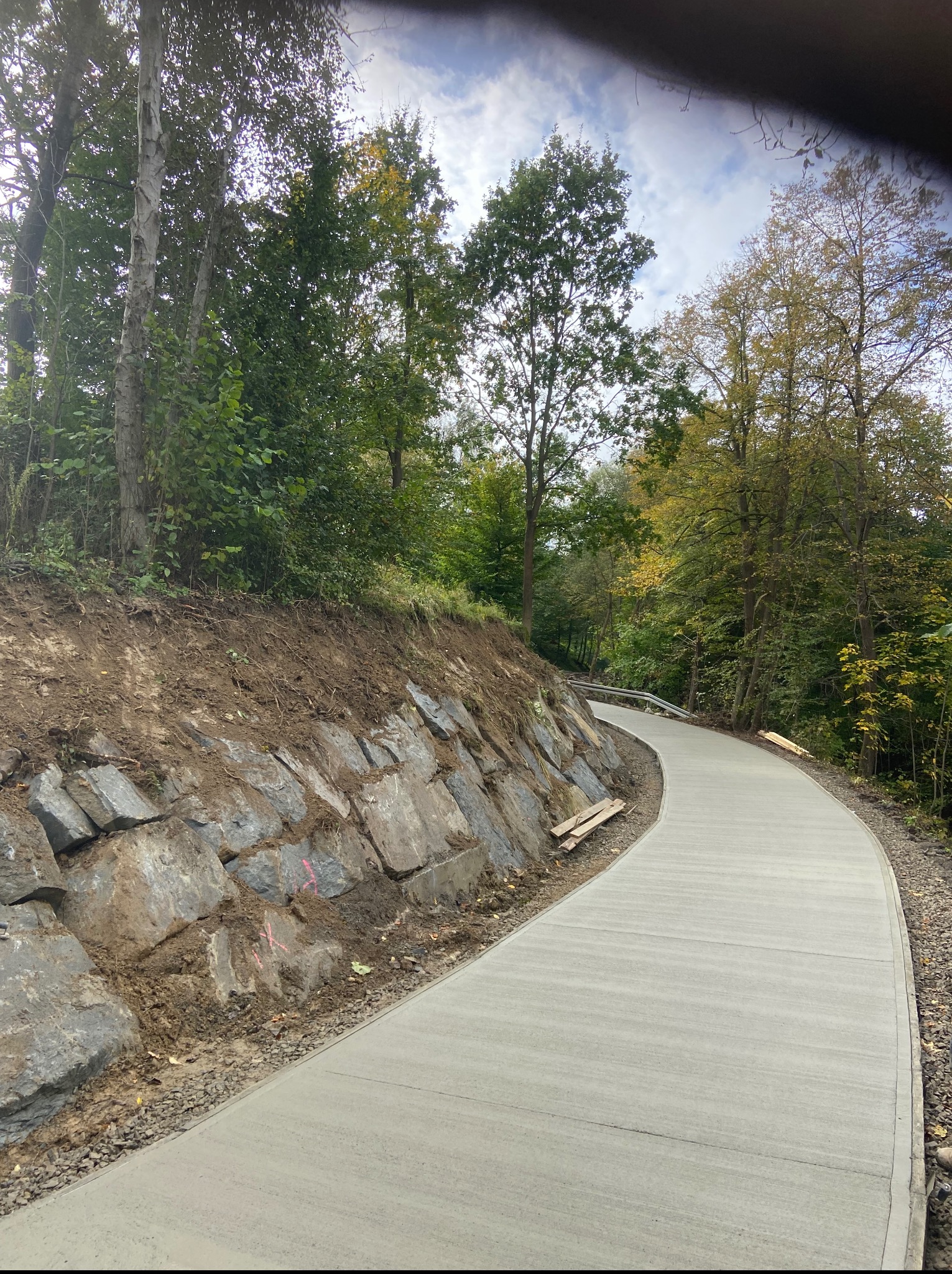 Nowa, betonowa ścieżka wijąca się przez zielony teren, obok kamiennego muru oporowego. Widoczne drzewa i fragment nieba w tle.
