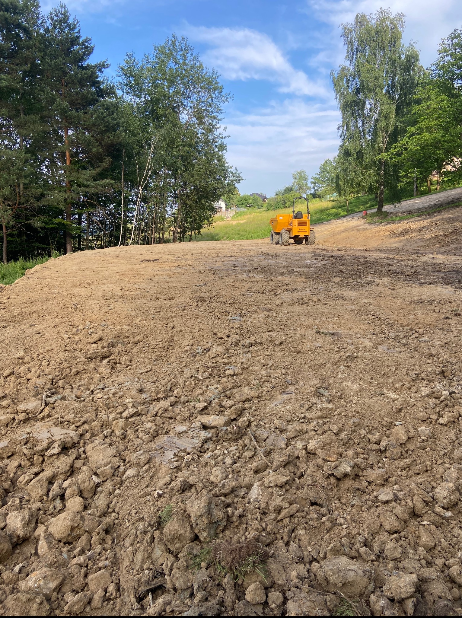 Żółta wywrotka na wyrównanym, brązowym terenie. W tle drzewa i błękitne niebo. Teren przygotowany pod dalsze prace budowlane, krajobraz po niwelacji.