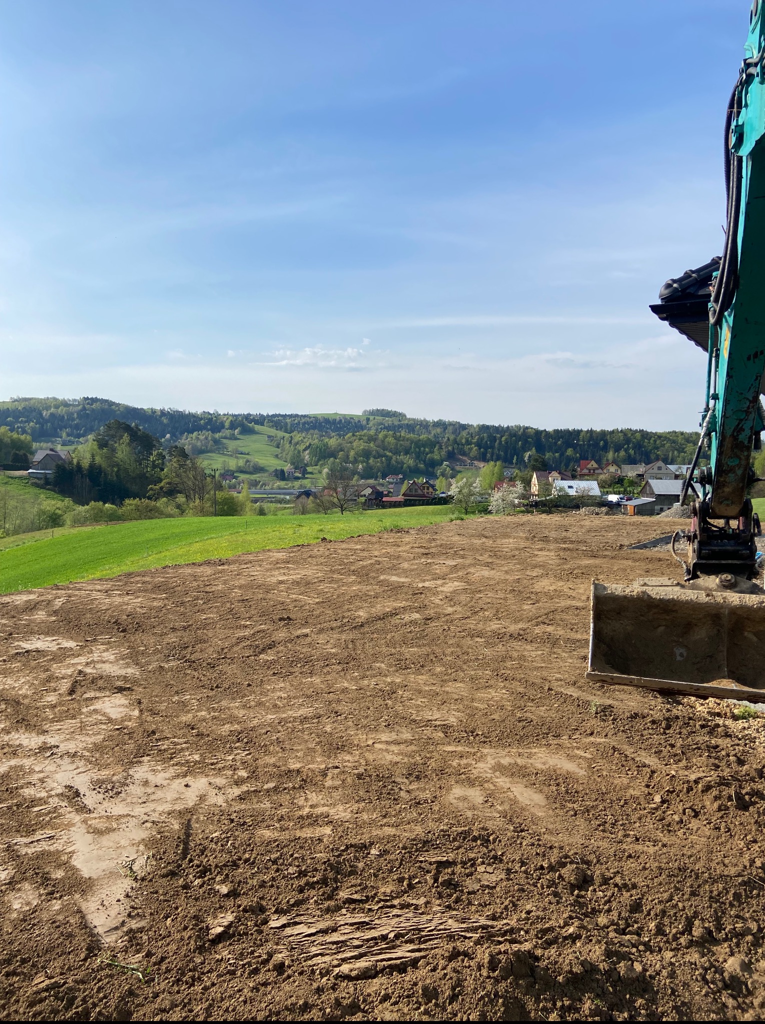 Niwelacja terenu w Bartkowej-Posadowej. Ziemia wyrównana, widoczny fragment koparki i zielone wzgórza w tle, błękitne niebo. Prace ziemne.