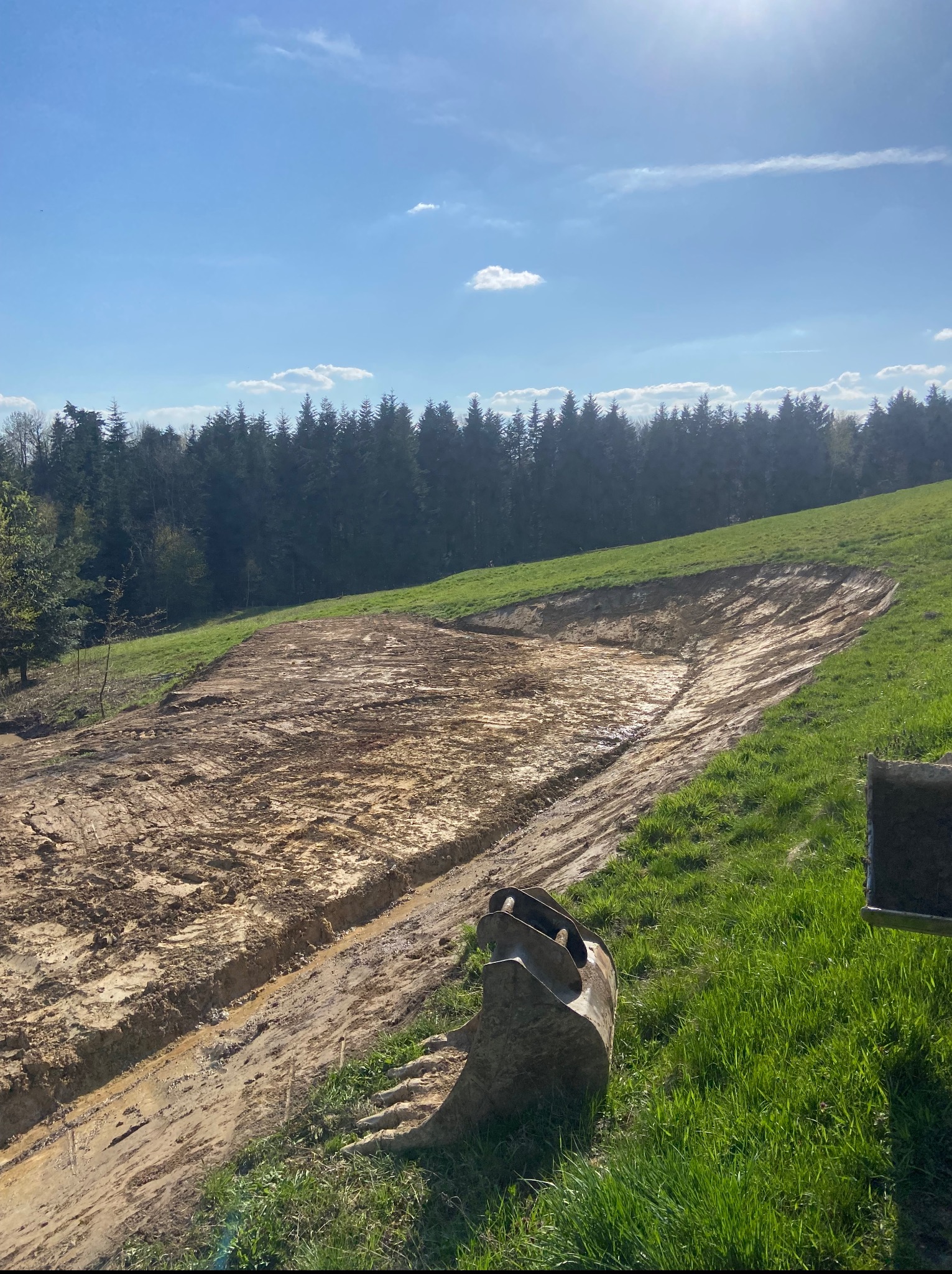 Wykopany teren pod staw na wzgórzu, z widocznym fragmentem łyżki koparki. Błękitne niebo i las w tle. Ślady pracy maszyn ziemnych na zboczu.