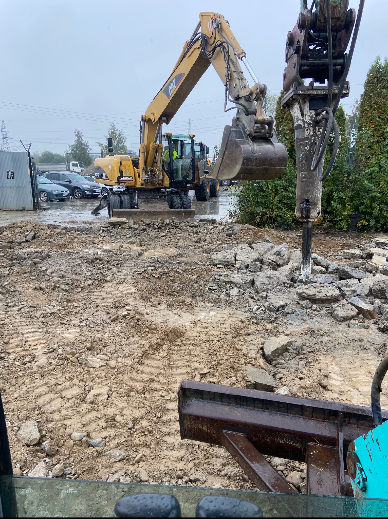 Żółta koparka CAT z łyżką i młotem hydraulicznym w trakcie rozbiórki gruzu na placu budowy. Widok z kabiny operatora, mokra nawierzchnia, pochmurne niebo.