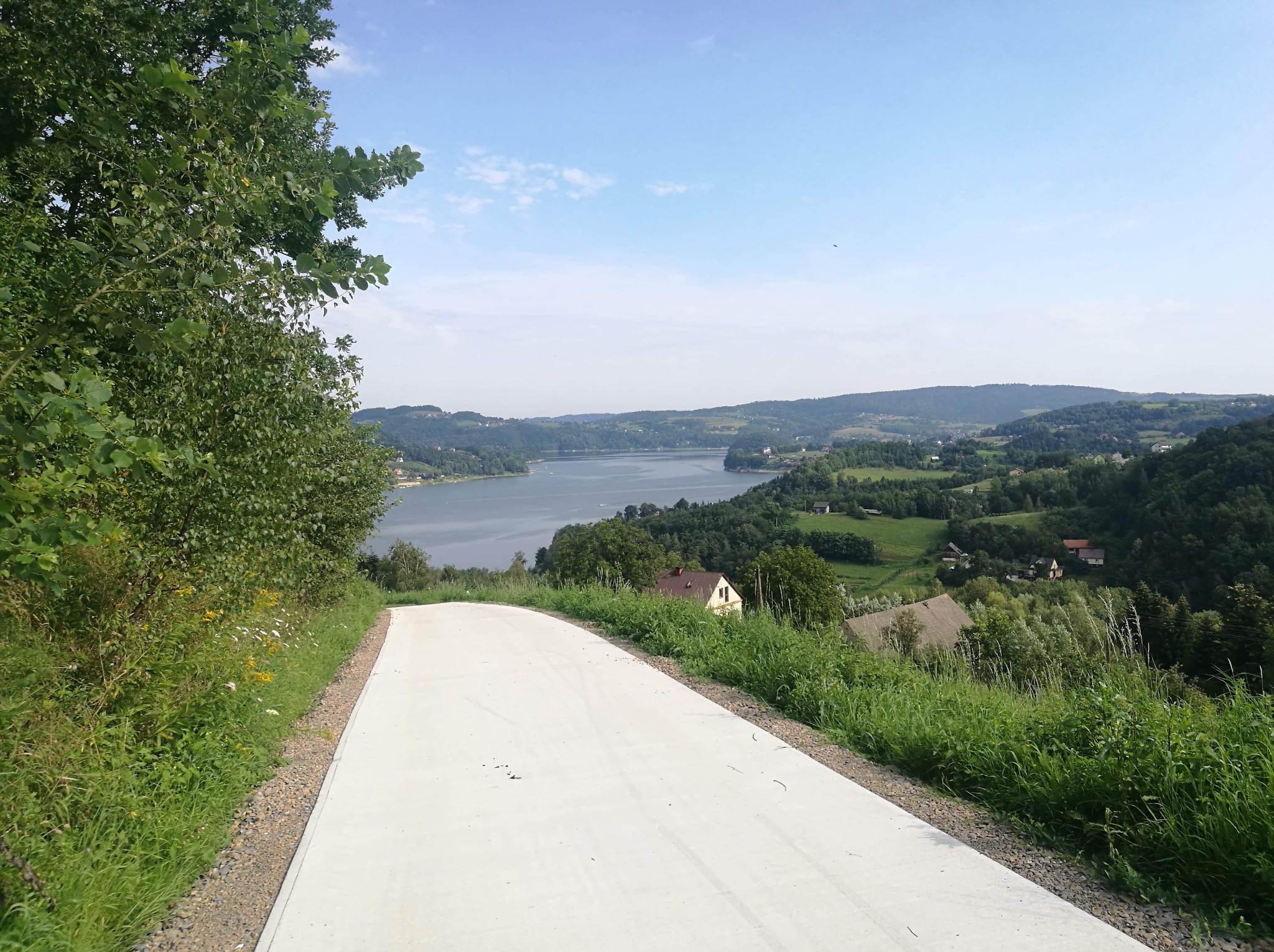 Betonowa droga prowadząca w dół wzgórza, z widokiem na malownicze jezioro i zielone wzgórza w tle. Widoczne elementy krajobrazu i roślinności.