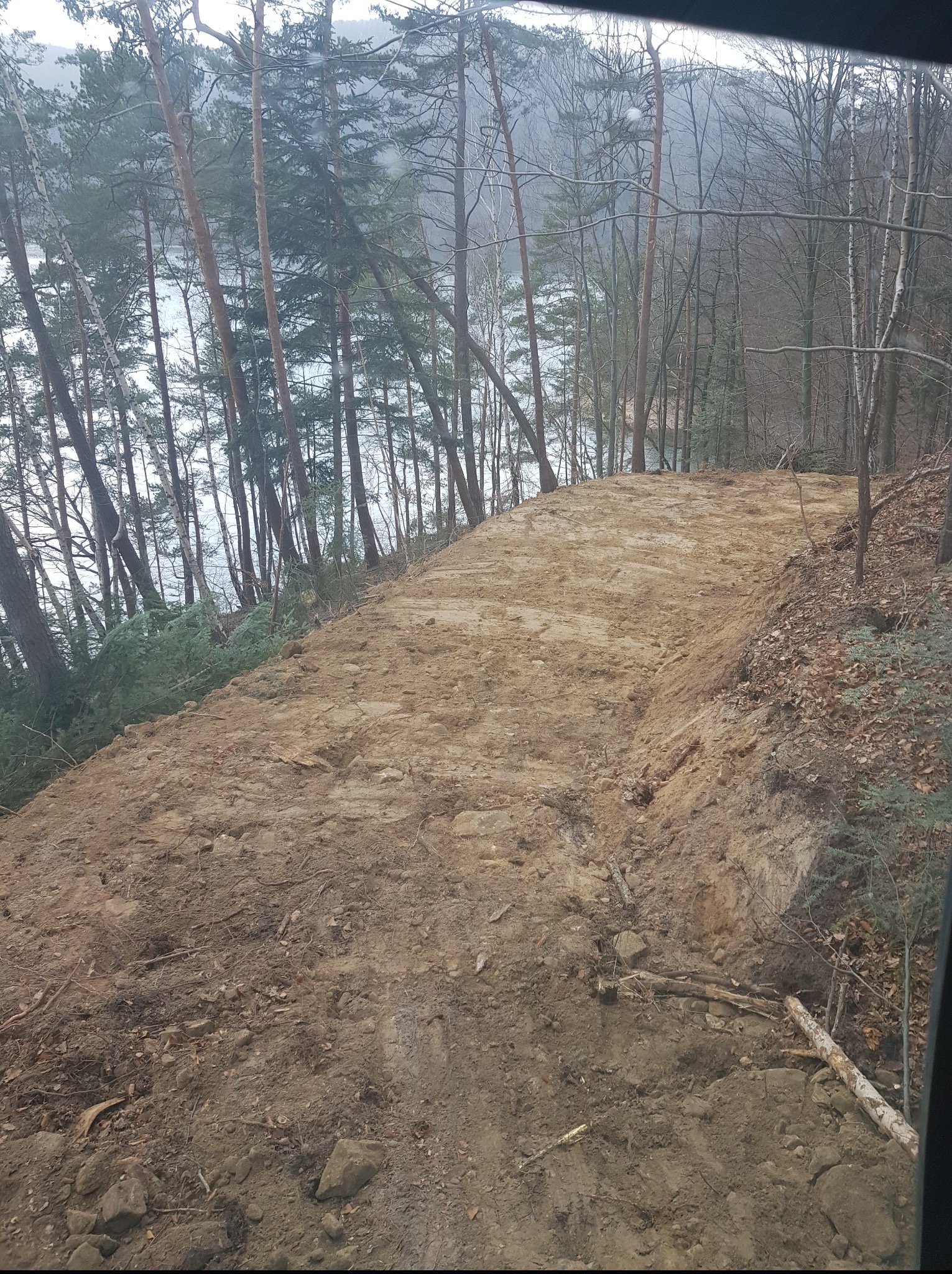 Ukształtowany teren pod przyszłą drogę lub ścieżkę, widok z góry. W tle drzewa i jezioro, pochmurna pogoda. Ślady opon na ziemi.