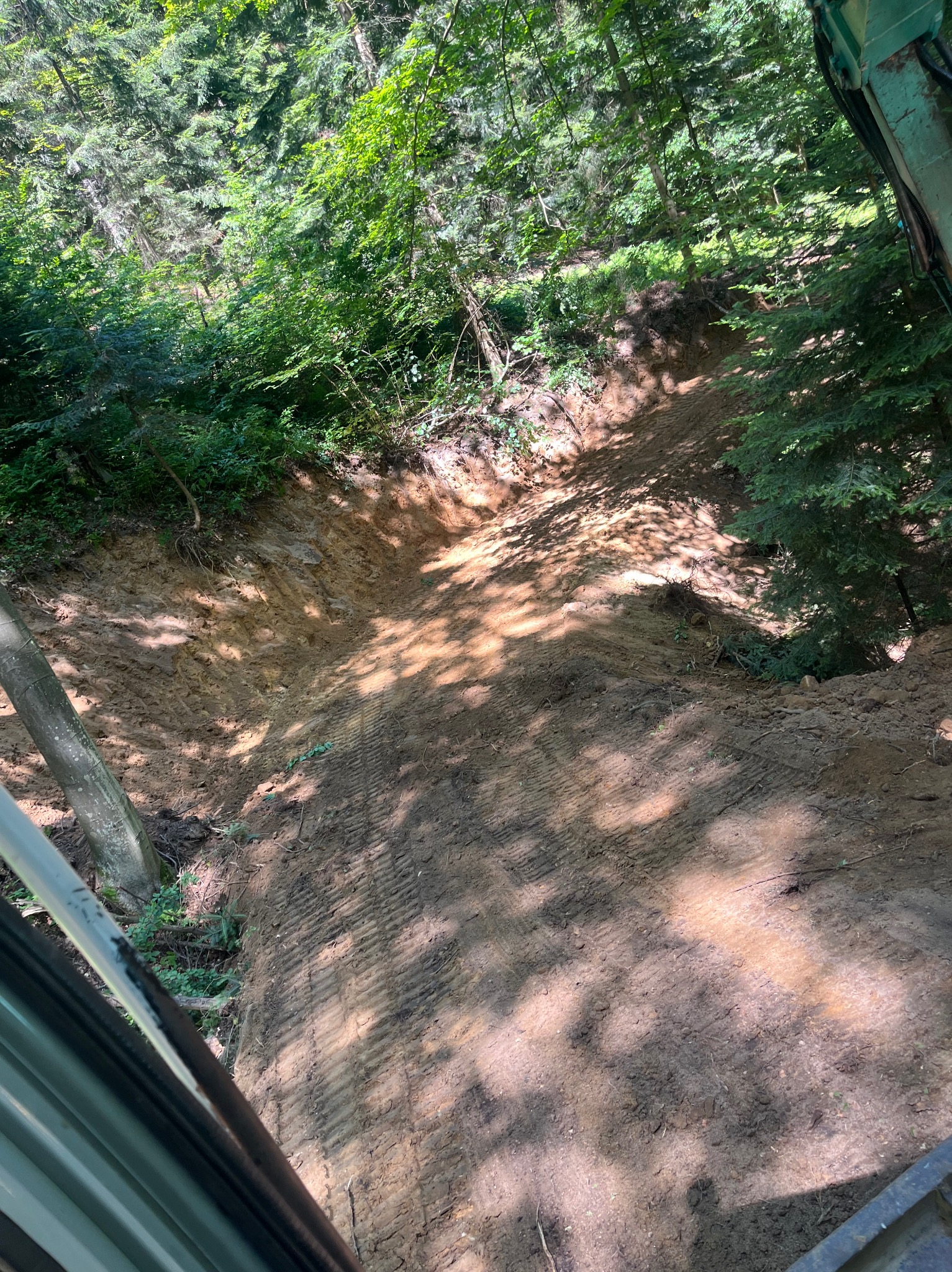 Świeżo wykopany teren w lesie, ślady opon, widok z kabiny maszyny. Teren wyrównany, przygotowany pod dalsze prace, w tle drzewa i krzewy.