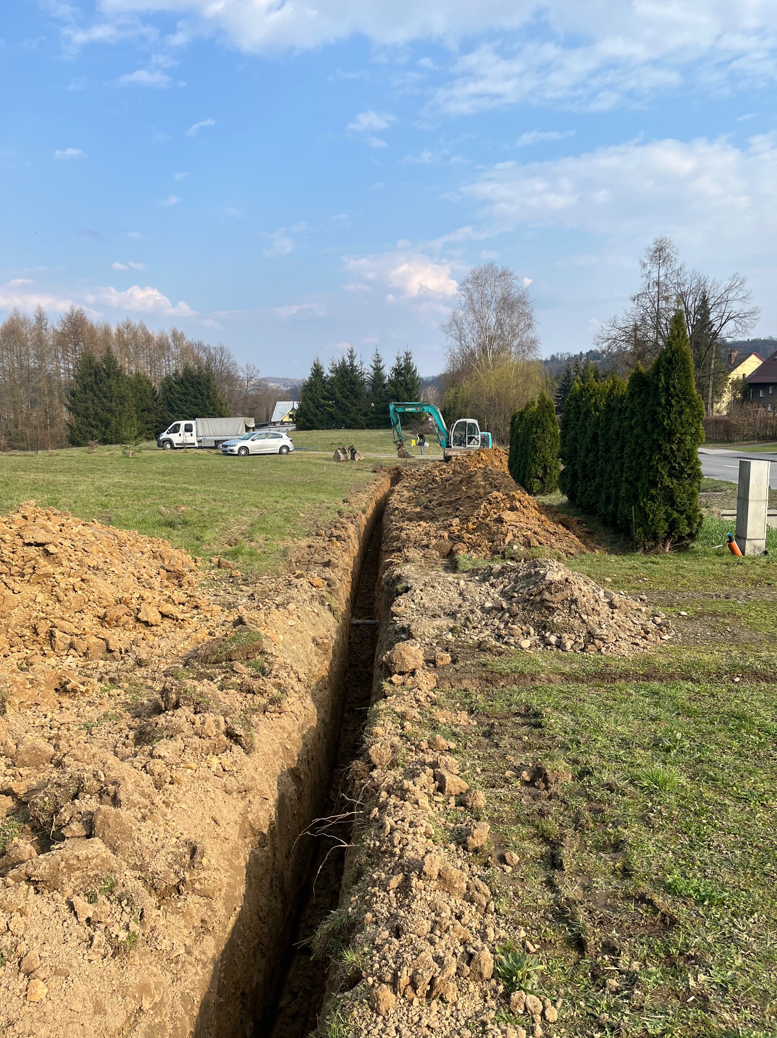 Wykopany wąski rów w ziemi na trawiastym terenie, z małą koparką w tle i stertami ziemi po bokach. Widoczne drzewa i zabudowania w oddali, błękitne niebo.